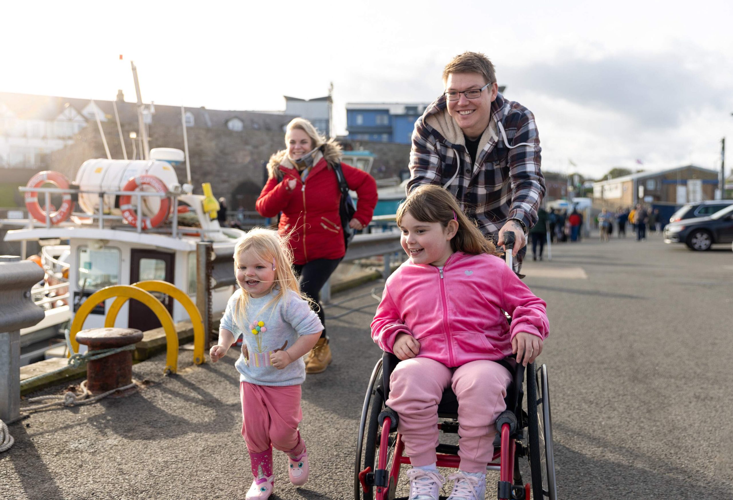 A family with two children enjoying a day out together in Beadnell, North East England. They are moving past the harbour and the main focus is the father running while pushing his daughter who is a wheelchair user. They all look excited.