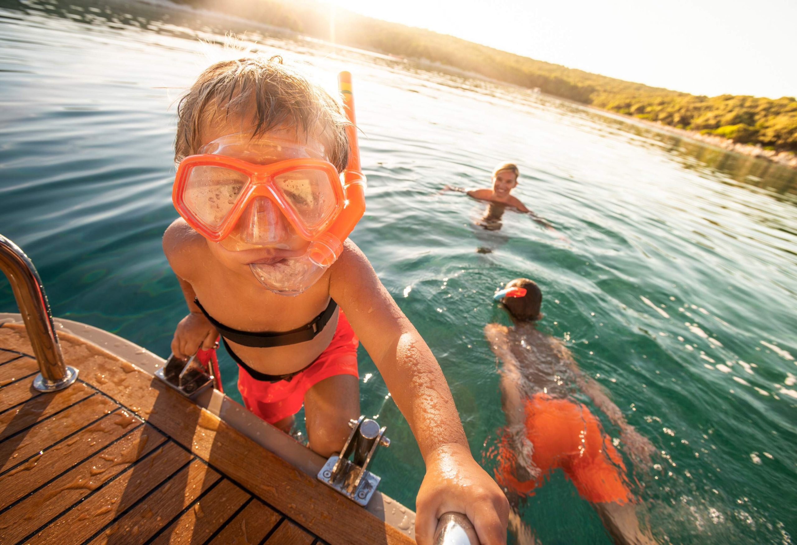 A little boy with a snorkel climbing a yacht ladder with two people in the water behind him.