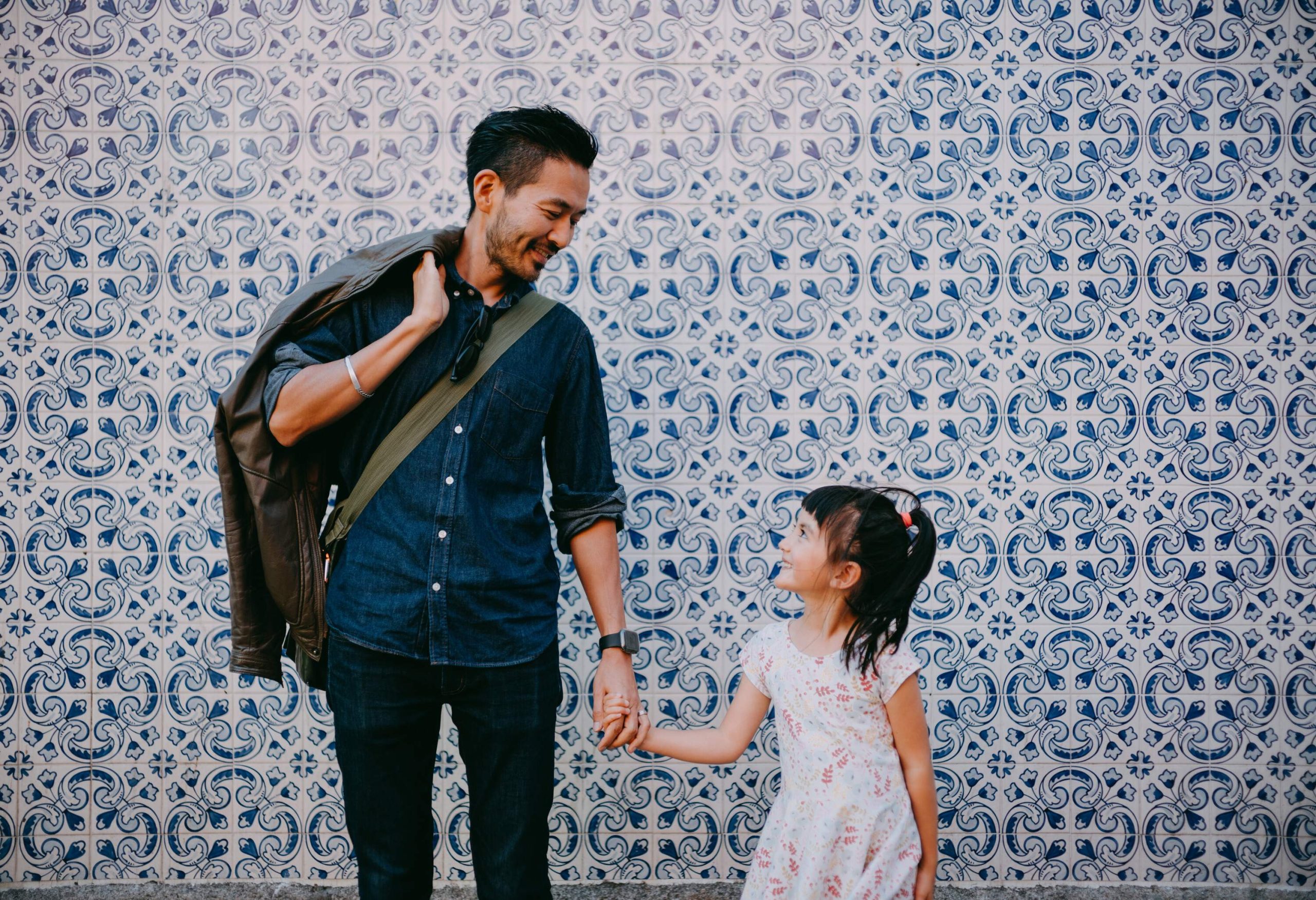A father standing against a white and blue tiled wall, making eye contact and holding his daughter's hand.