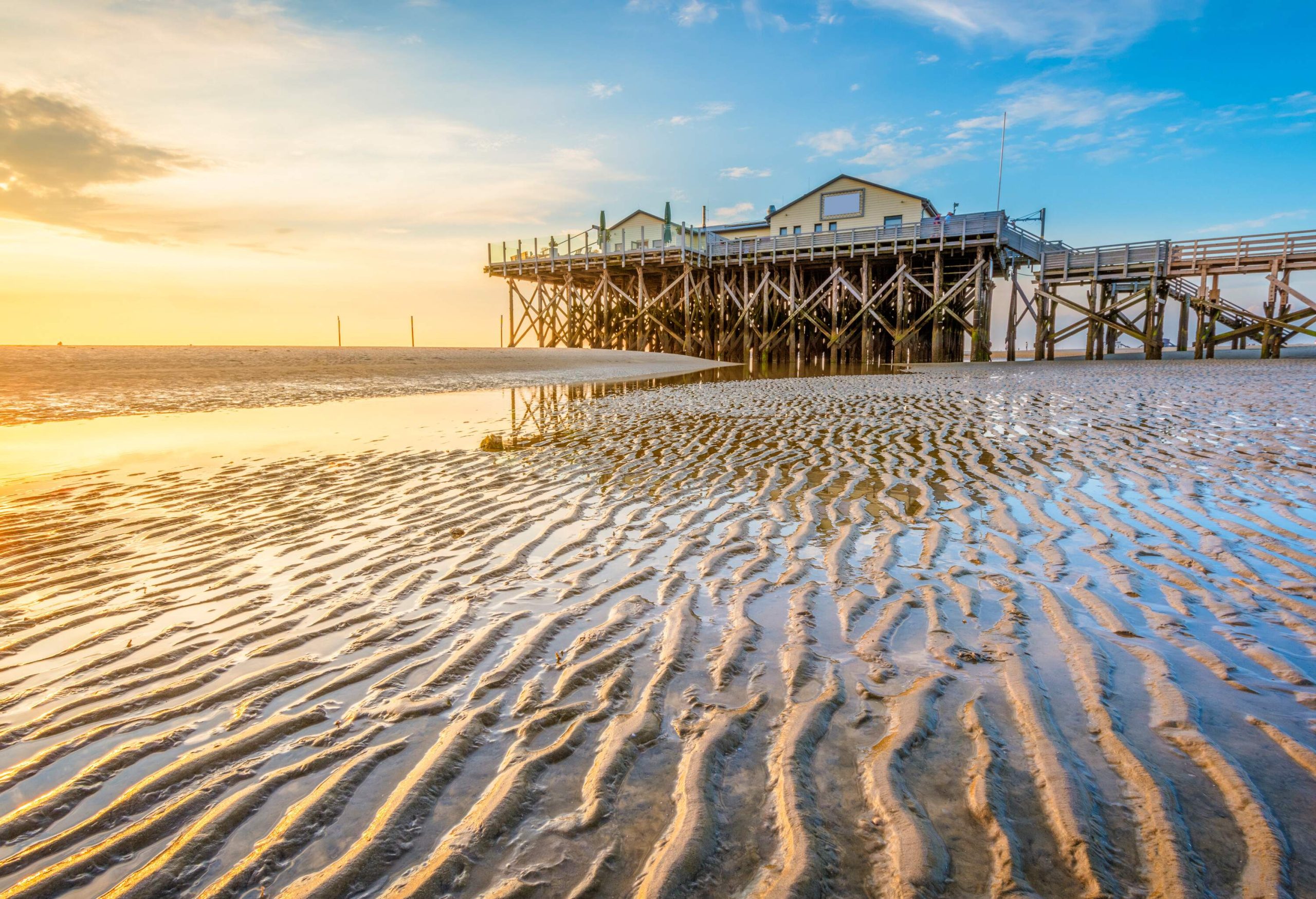 A stilt house perched above a beach with sand wave patterns at sunset.