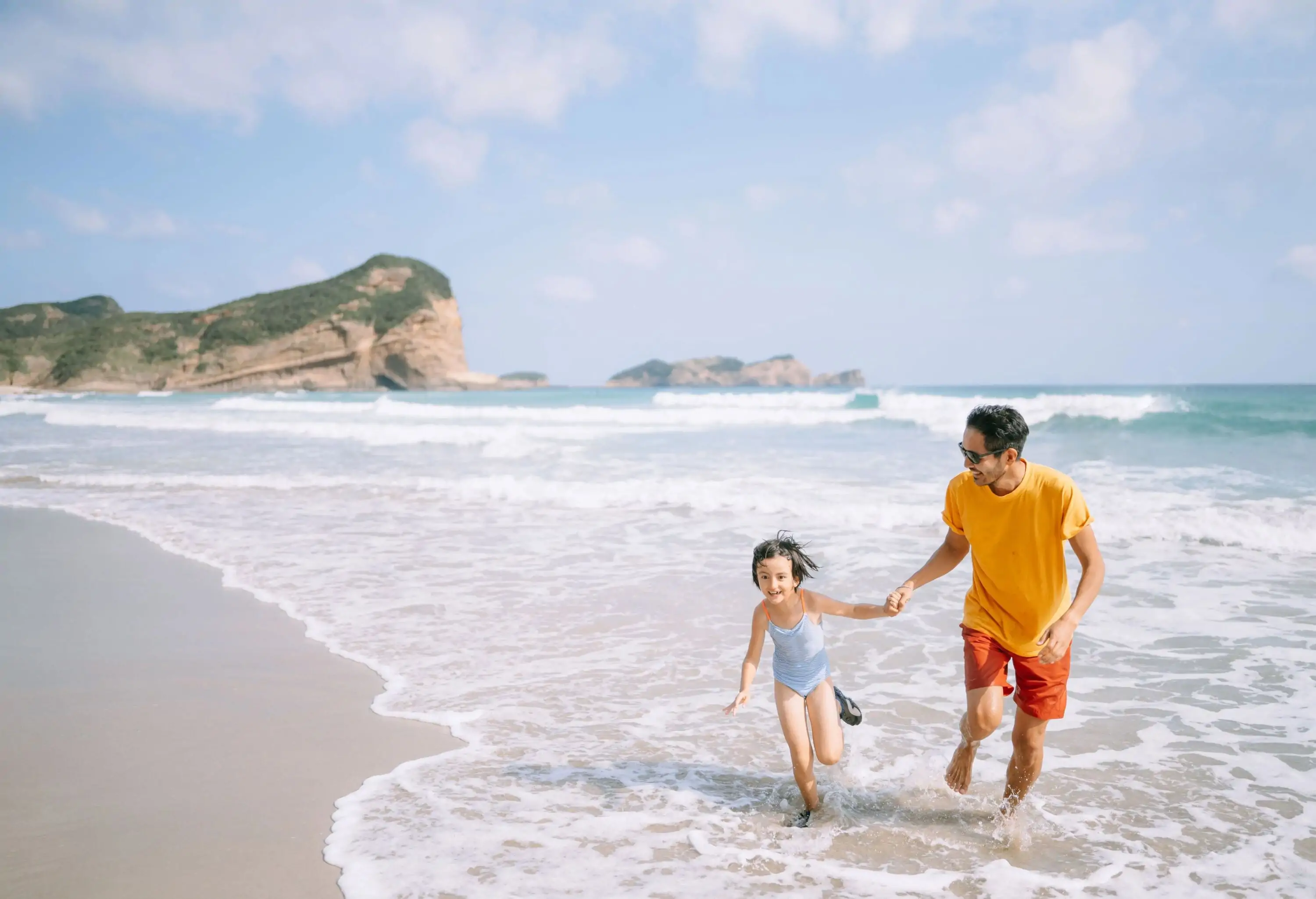 A father holds his daughter's hand while running on the beach.