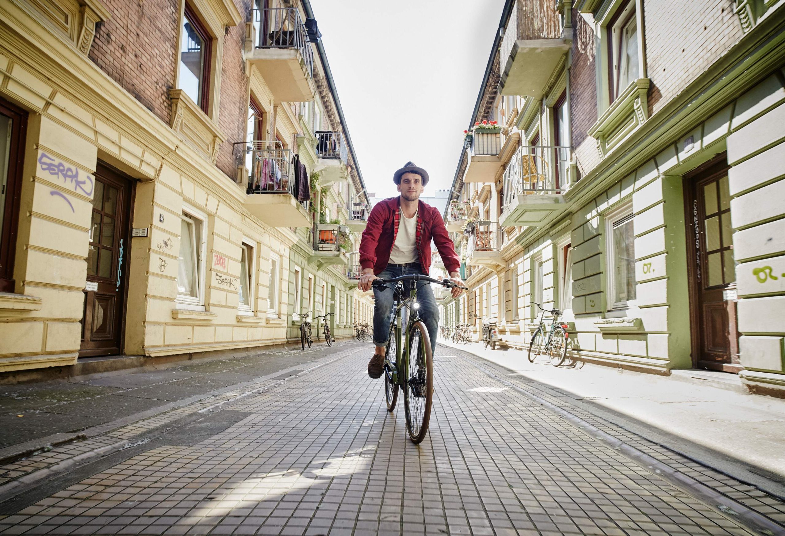 A man in a brown sweater and hat rides a bicycle as he travels on a street bordered by adjacent apartment buildings.