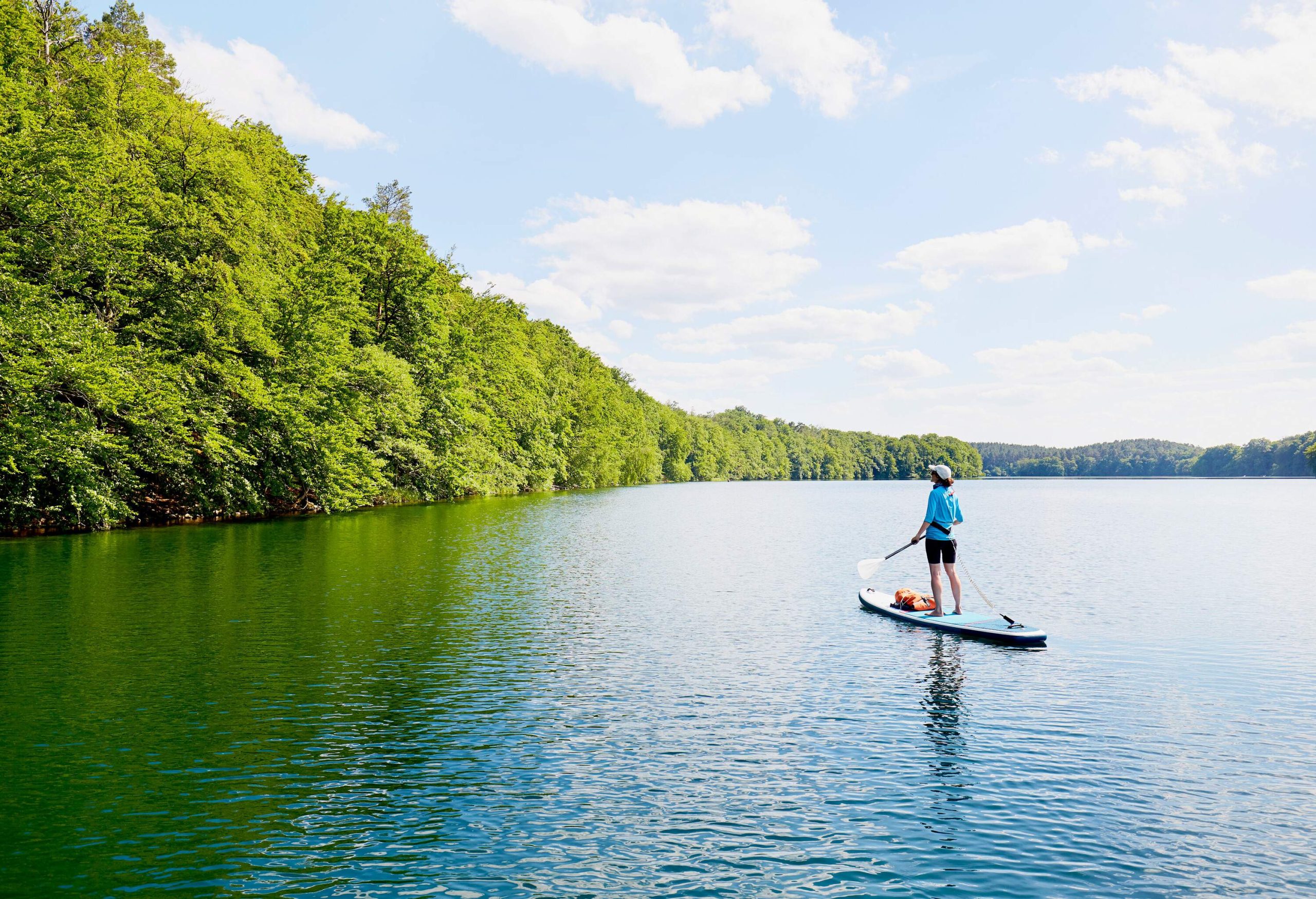 A woman paddleboarding across a calm lake circled by densely forested hills.