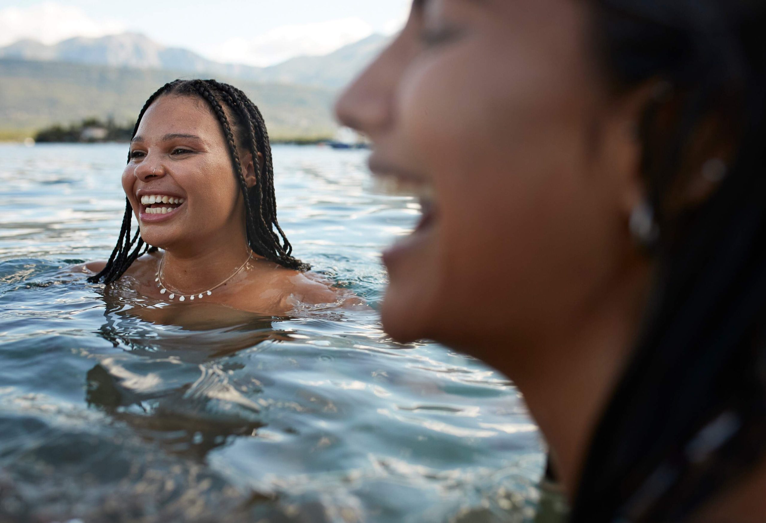 A woman with braided hair laughs with her friend while immersed in water.