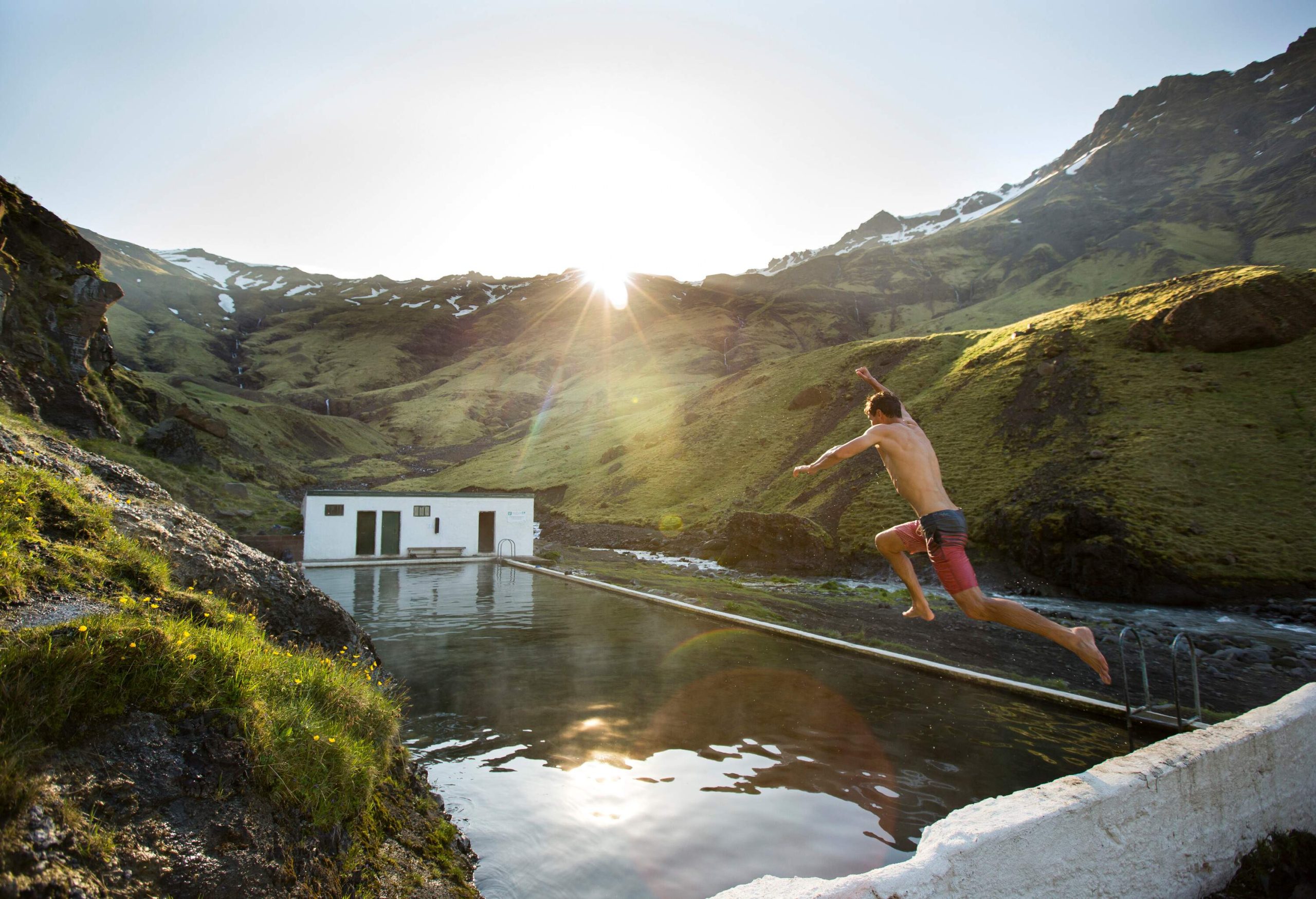 A man jumps into a geothermal hot spring, surrounded by rugged hills with sparse vegetation and a white structure visible on the other side of the pool.