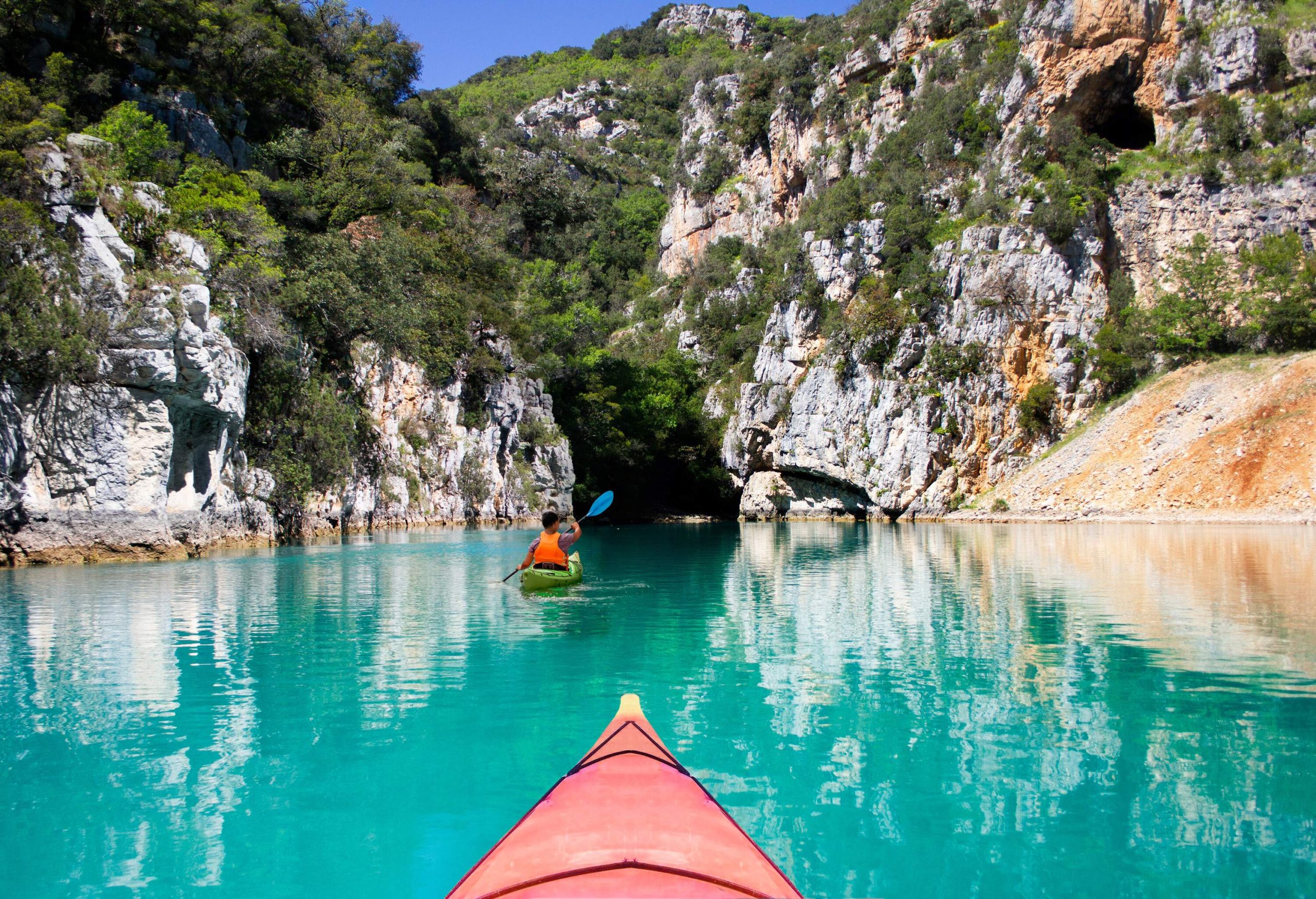 A group of kayakers paddles across a tranquil turquoise river in a gorge covered in vegetation.