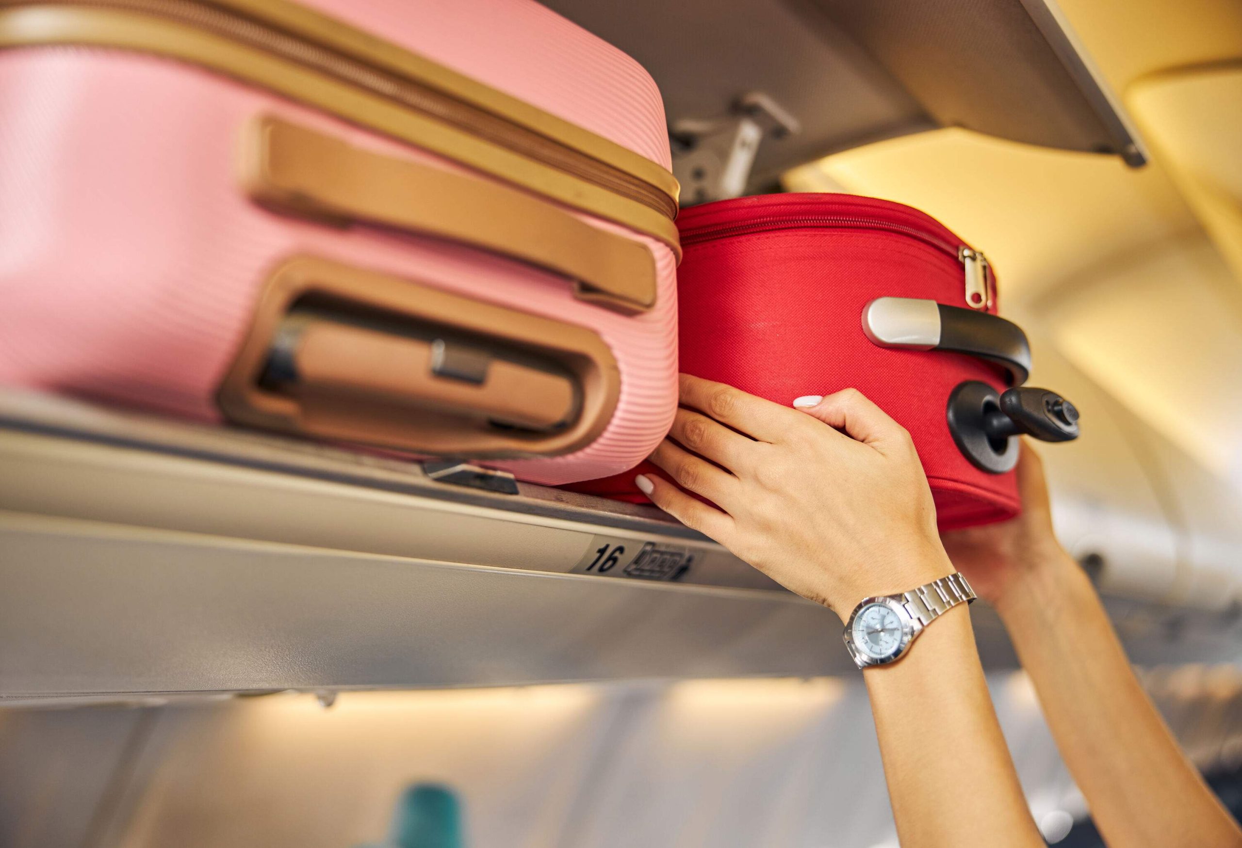 A woman squeezing a red suitcase into the overhead compartment's limited space.