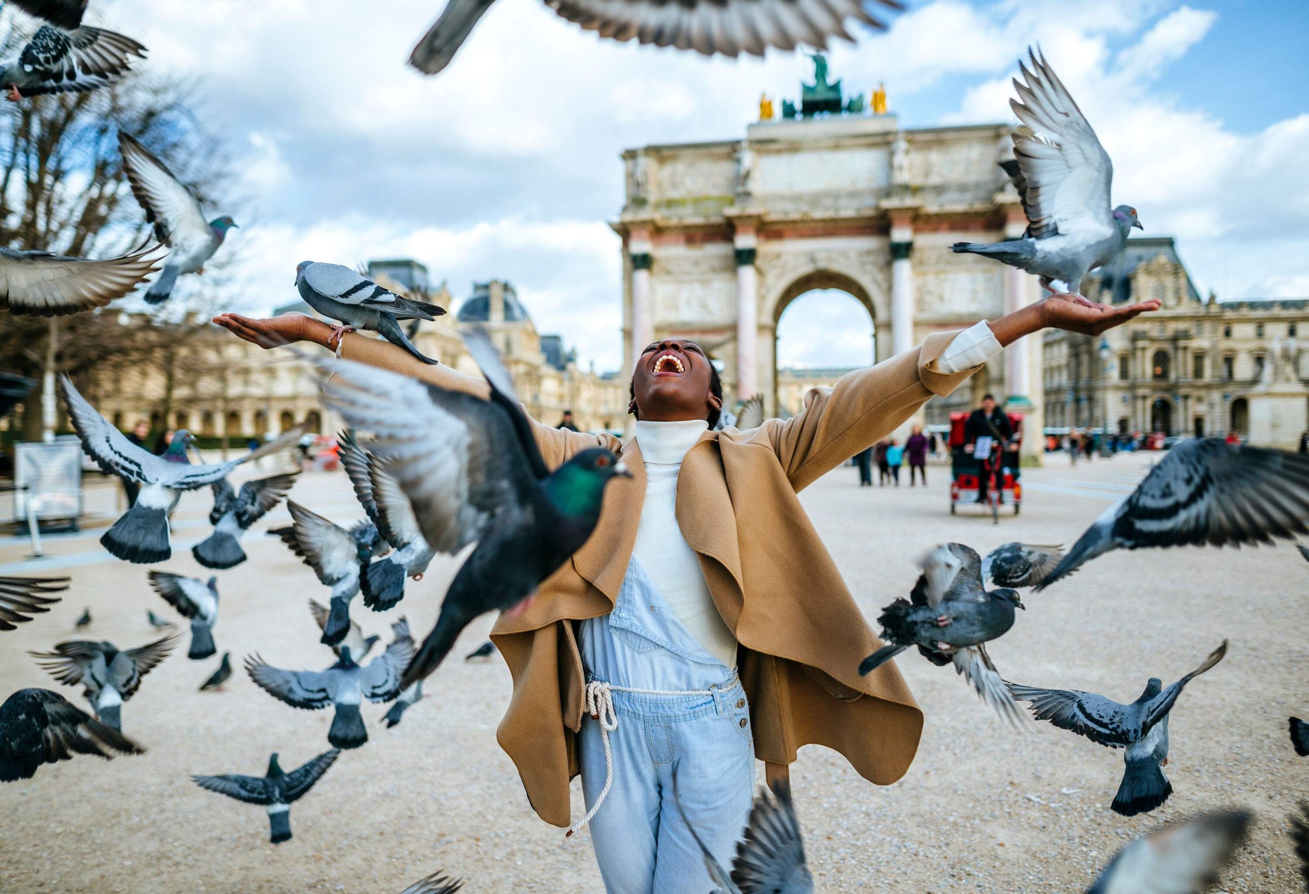 A joyful woman raises her hands in delight as she is surrounded by a flock of flying doves on the street, with the iconic Arc de Triomphe in Paris serving as a majestic backdrop.