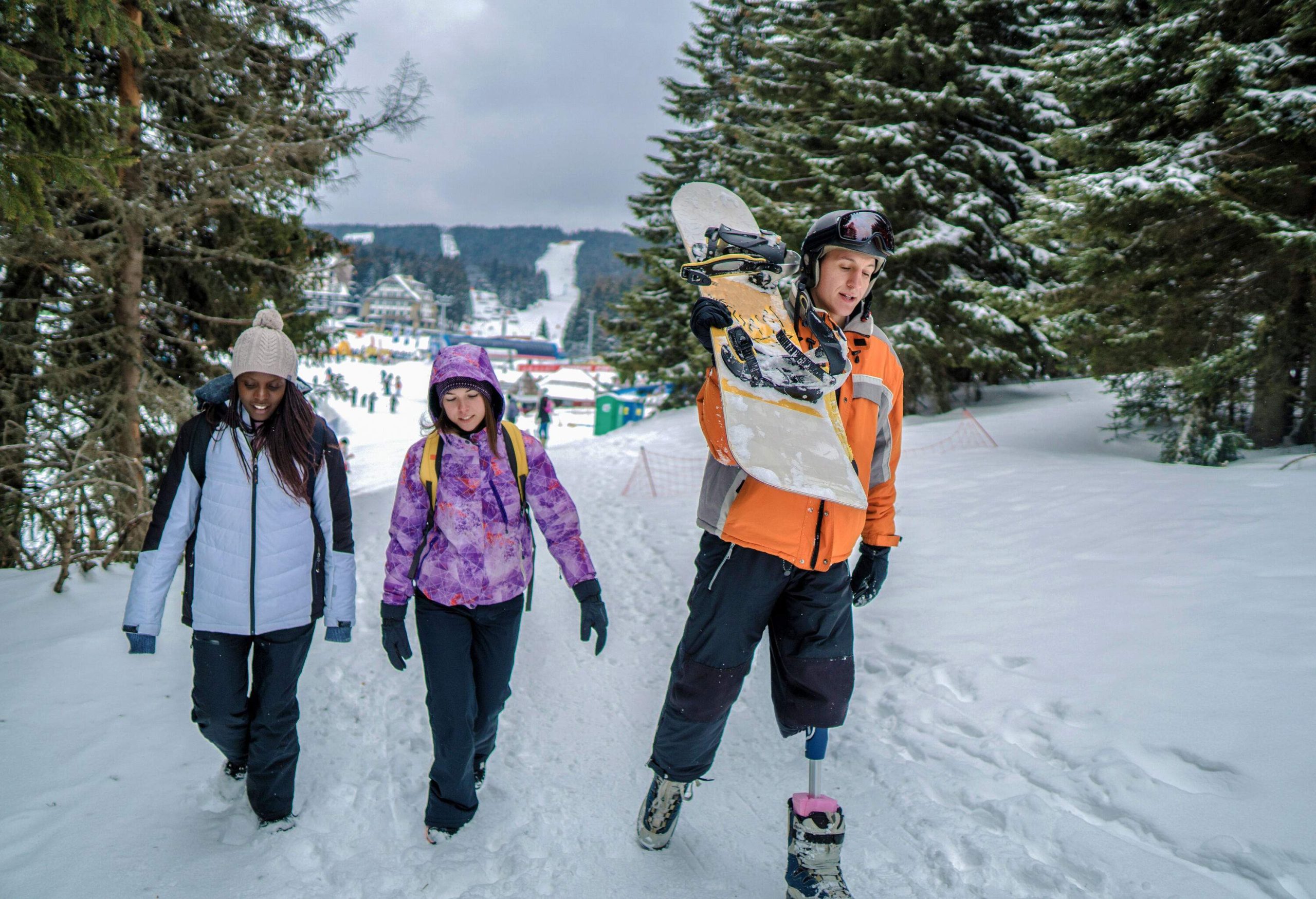 Man with prosthetic legs strolling alongside two friends across a snowy area bordered by trees.