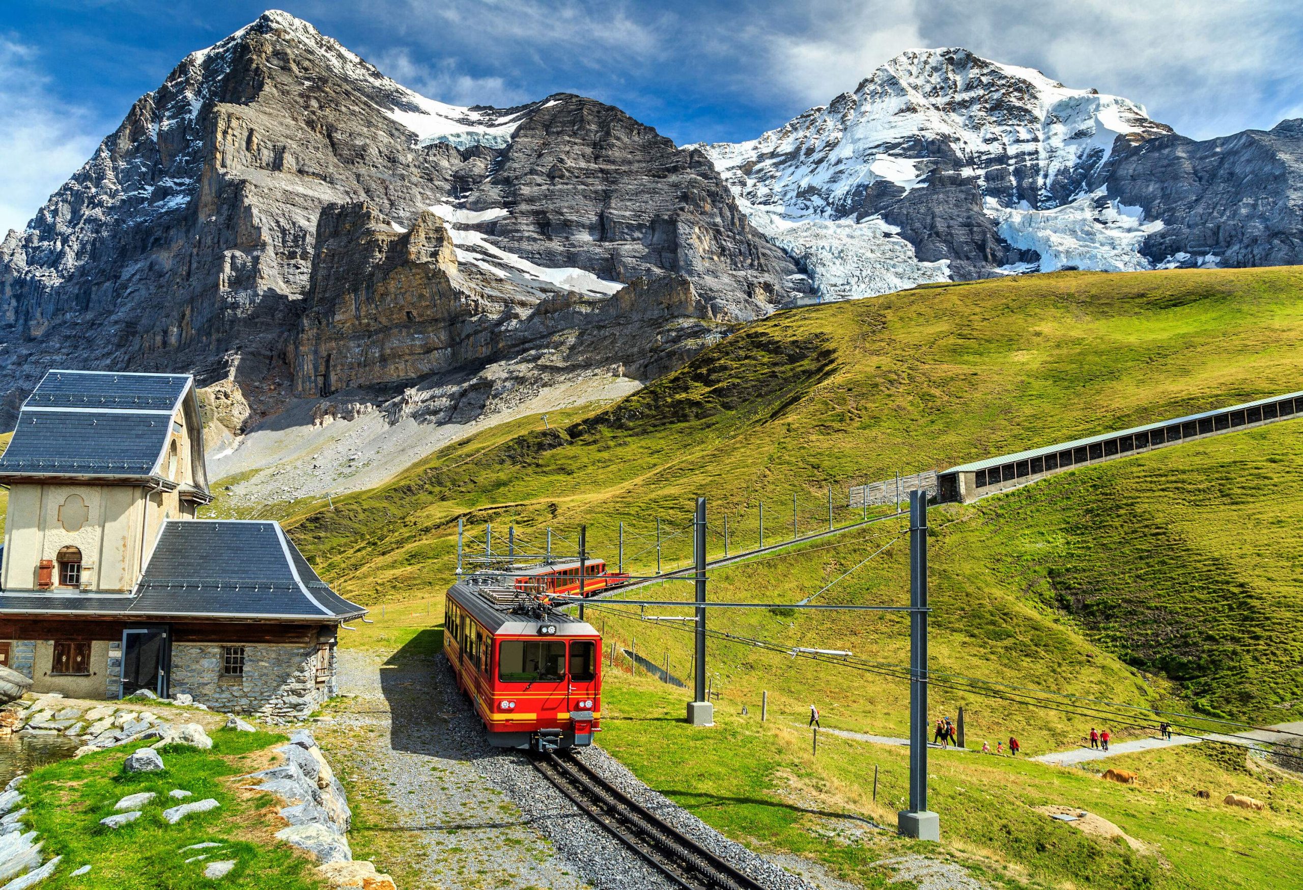An electric red tourist train winds its way through a verdant landscape, with the imposing presence of rugged mountains forming a dramatic backdrop in the distance.