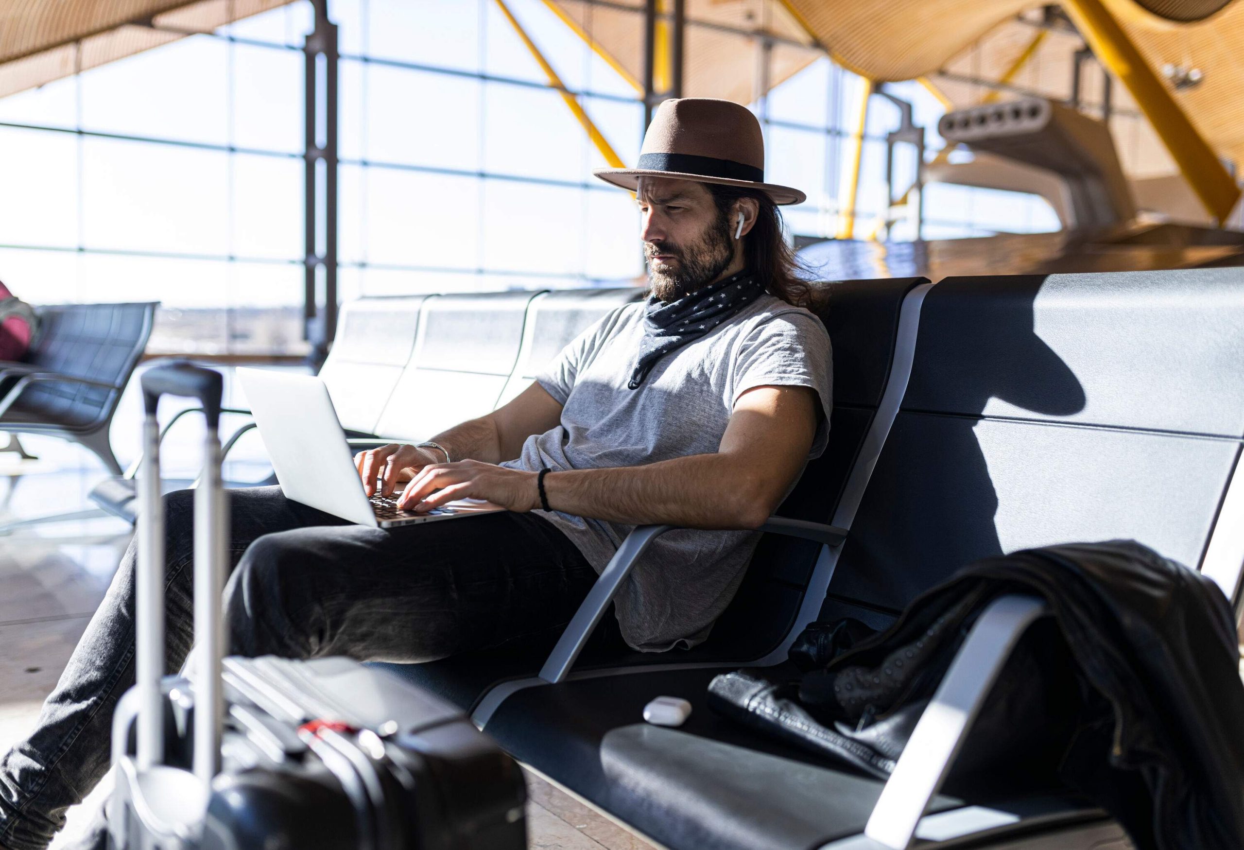 A man with a fedora hat and wireless earphones working on his laptop while slouched on one of the seats in a terminal.