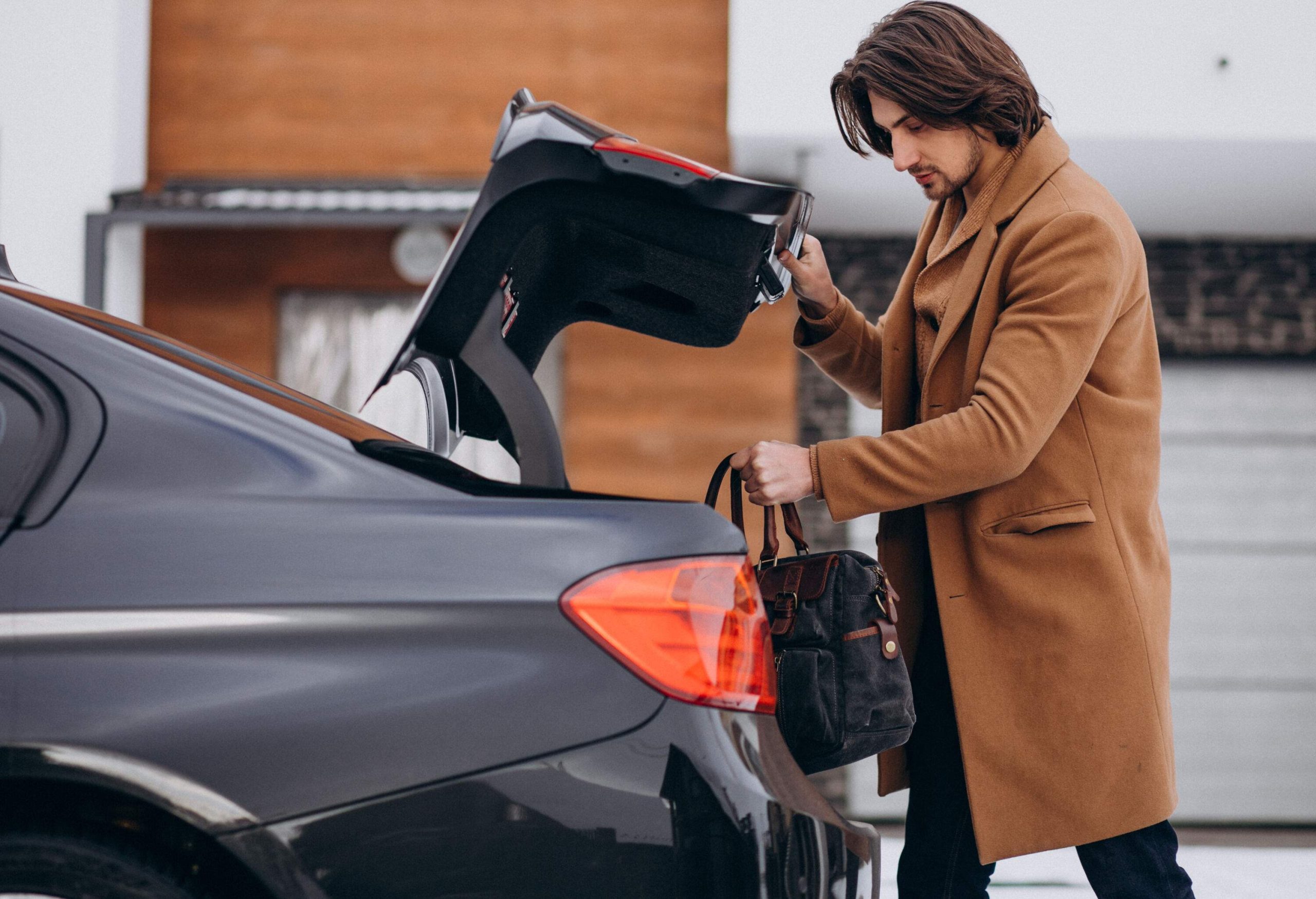 A man in a brown winter coat is putting his bag in the trunk of his car.