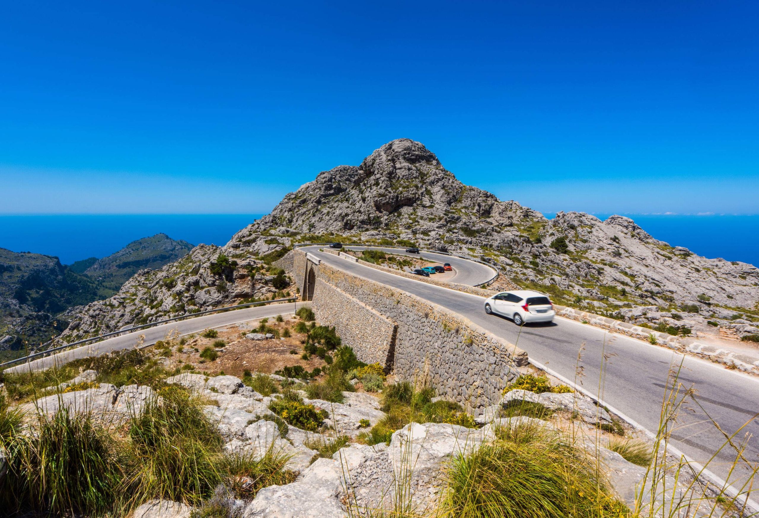Vehicles travel the spiral bridge road in the rocky mountain overlooking the sea against the blue sky.