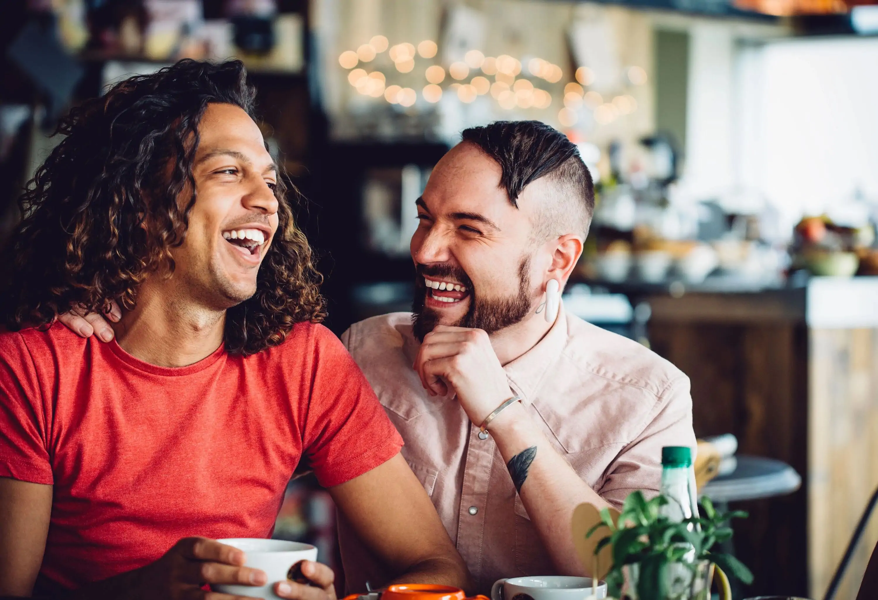 Two male friends laugh with each other while having coffee inside a café.