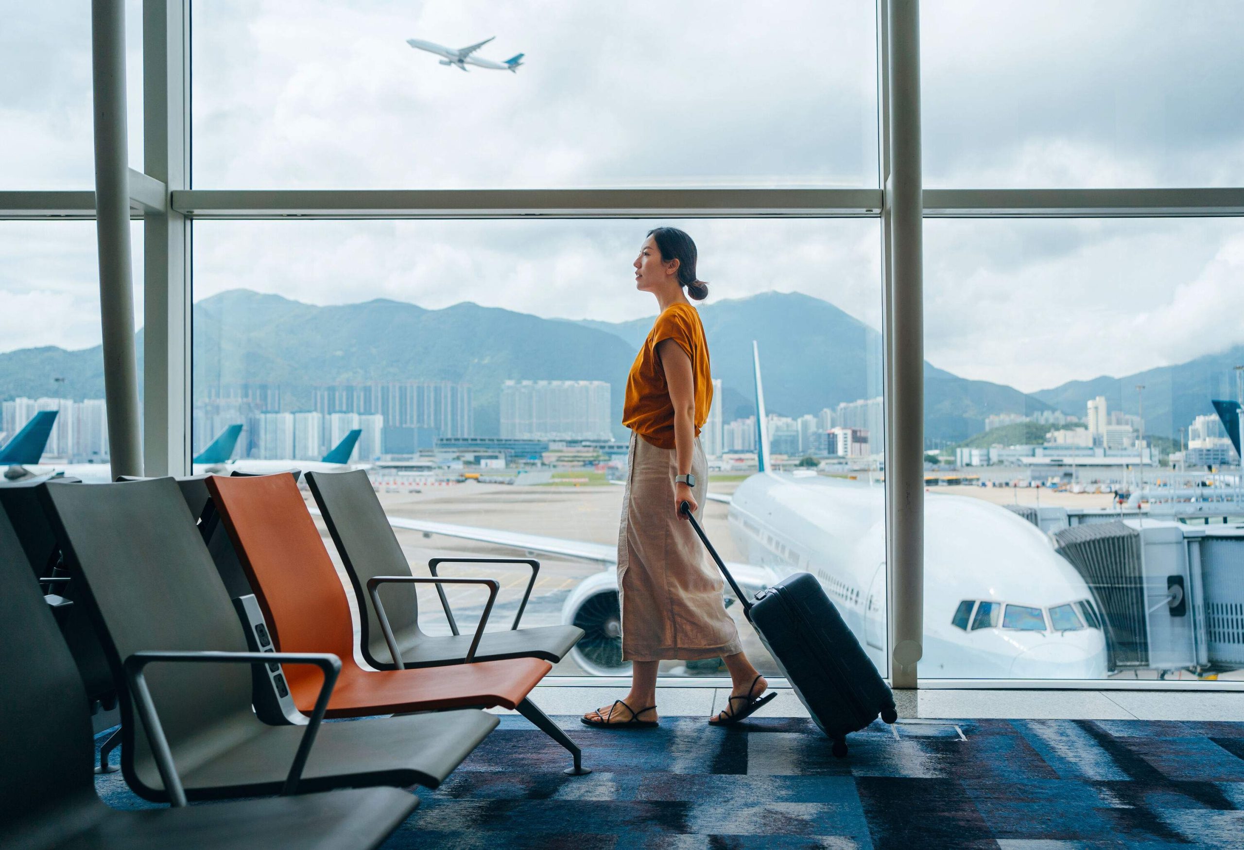 Young woman carrying suitcase, walking by the window at airport terminal. Young Asian female traveller waiting for boarding at airport. Business travel. Travel and vacation concept