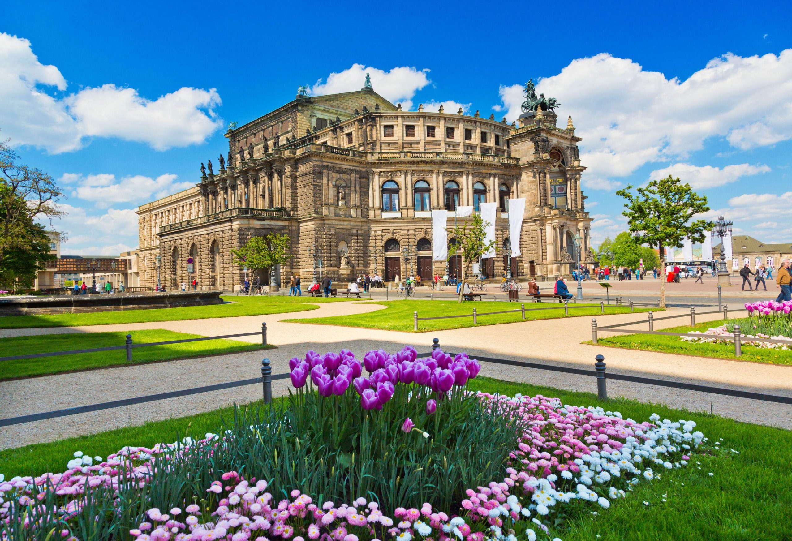 An opera house building fronting a square landscaped with colourful flowers, green grass, and trees with several people around.