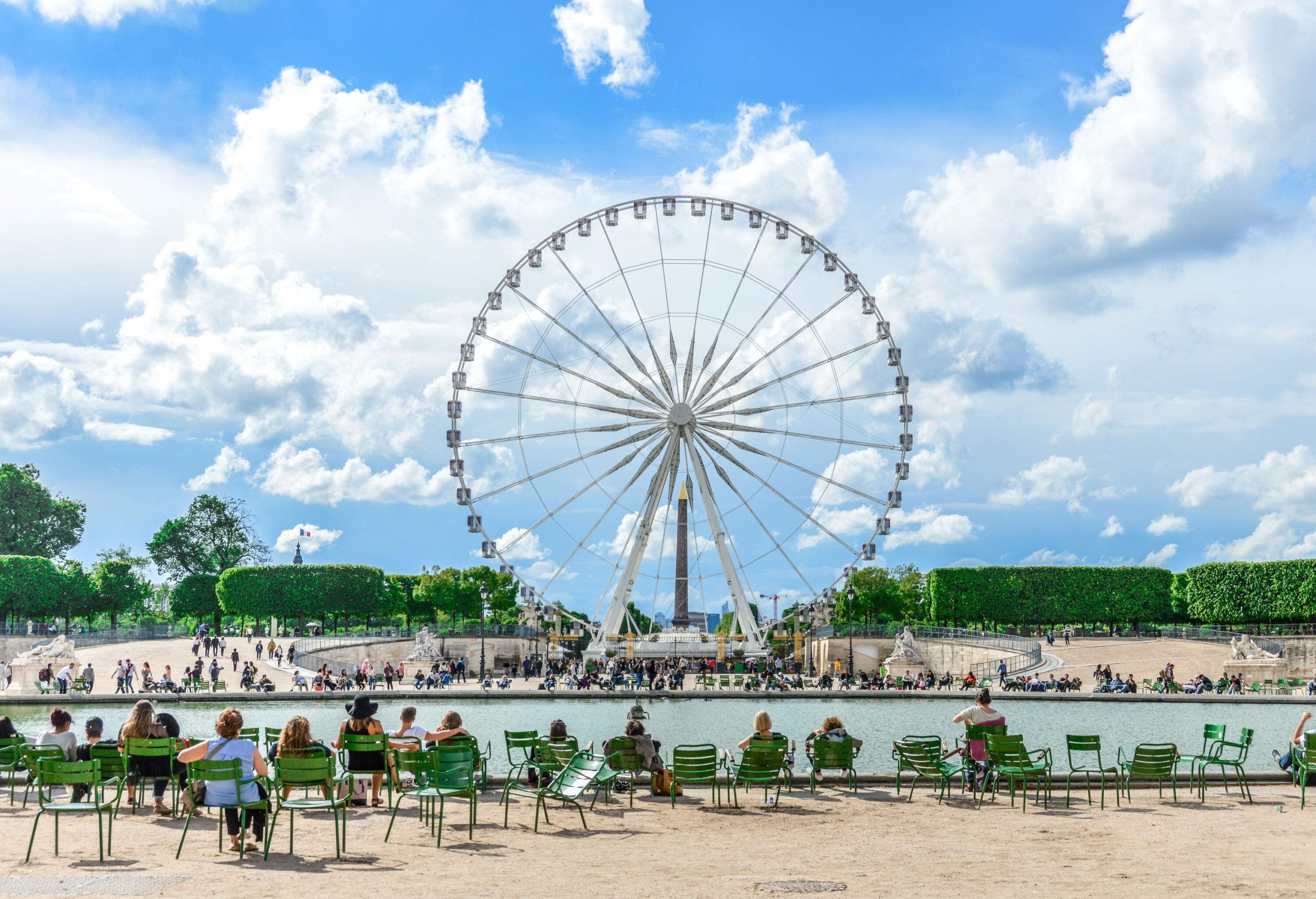 People flocked around a huge Ferris wheel alongside a pond surrounded by lush trees.