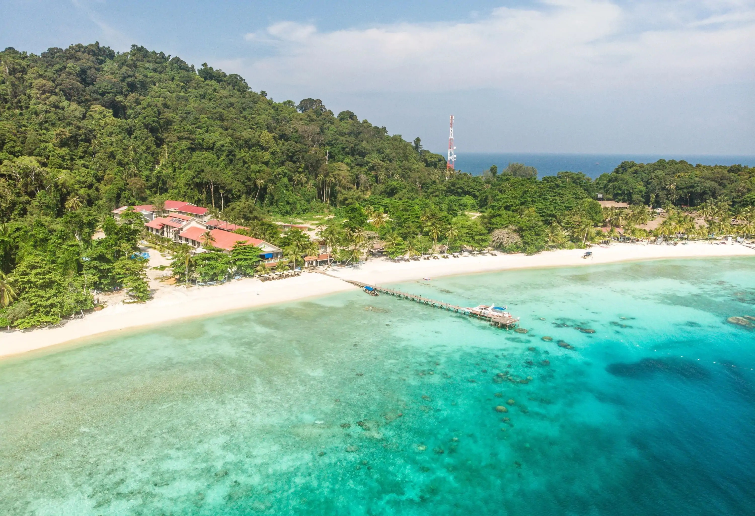 A beachfront resort at the base of a forested hill with a pier across the ocean with turquoise waters.