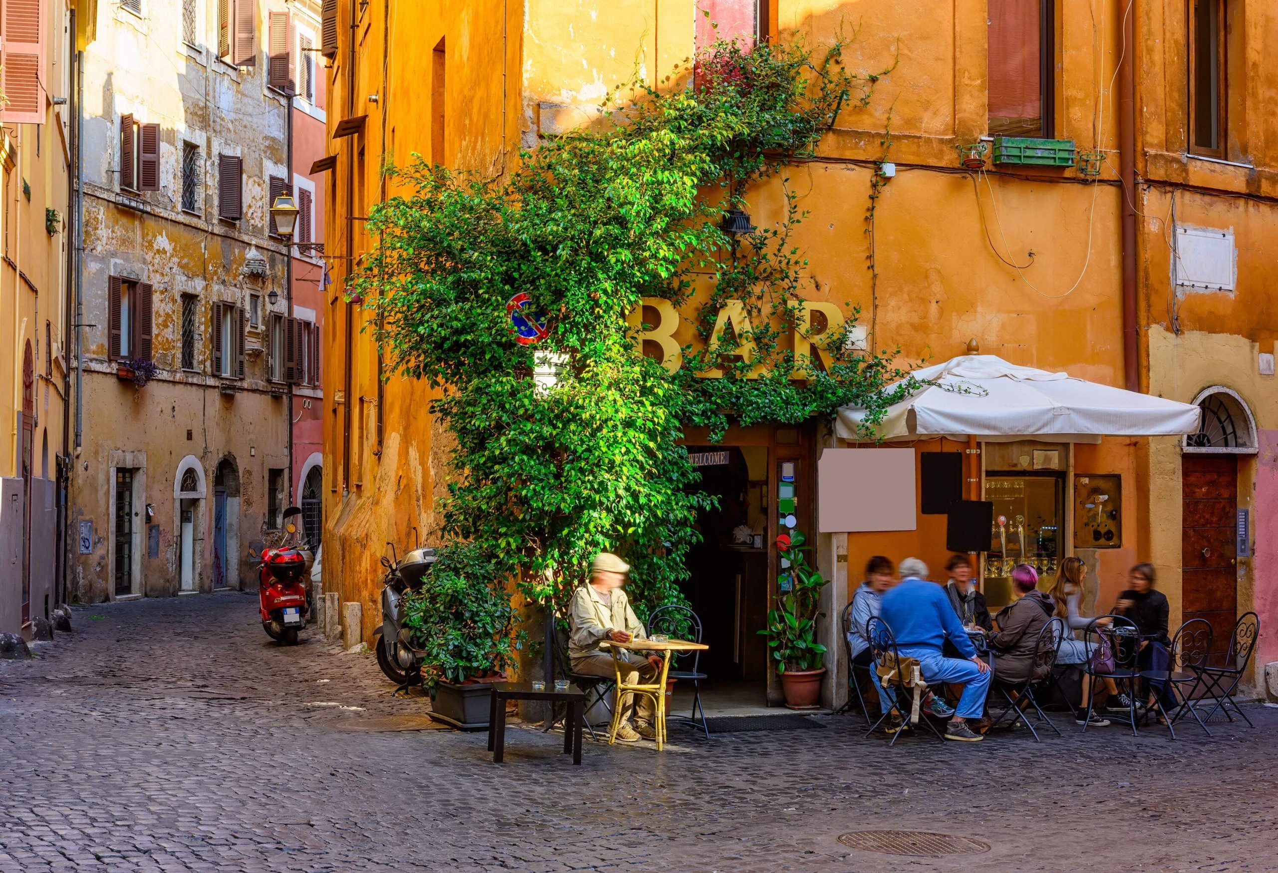 People socialise in an outdoor bar adjacent to a narrow street lined with traditional townhouses.