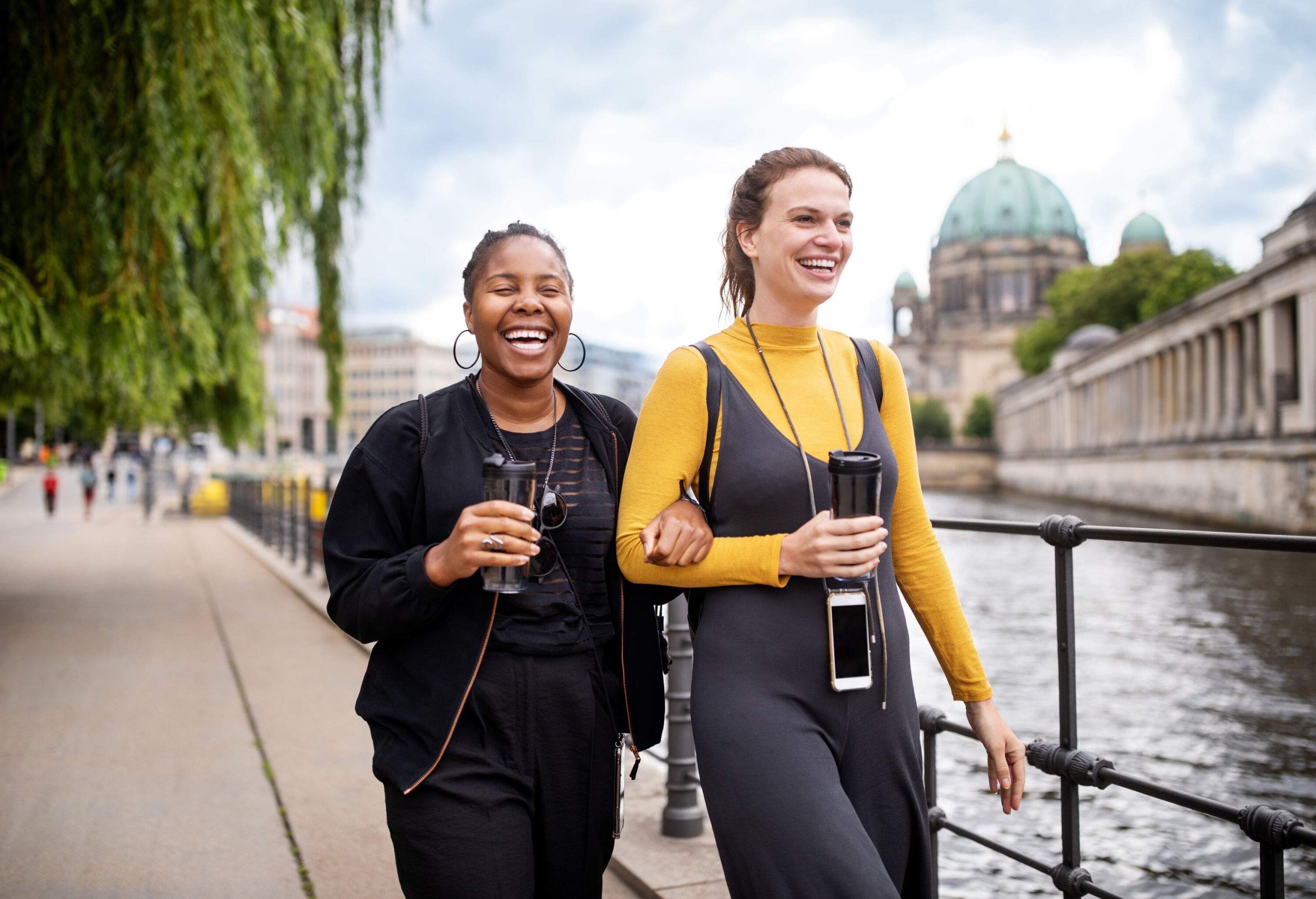 Two female friends in casual clothes smile as they walk down a promenade along a river.