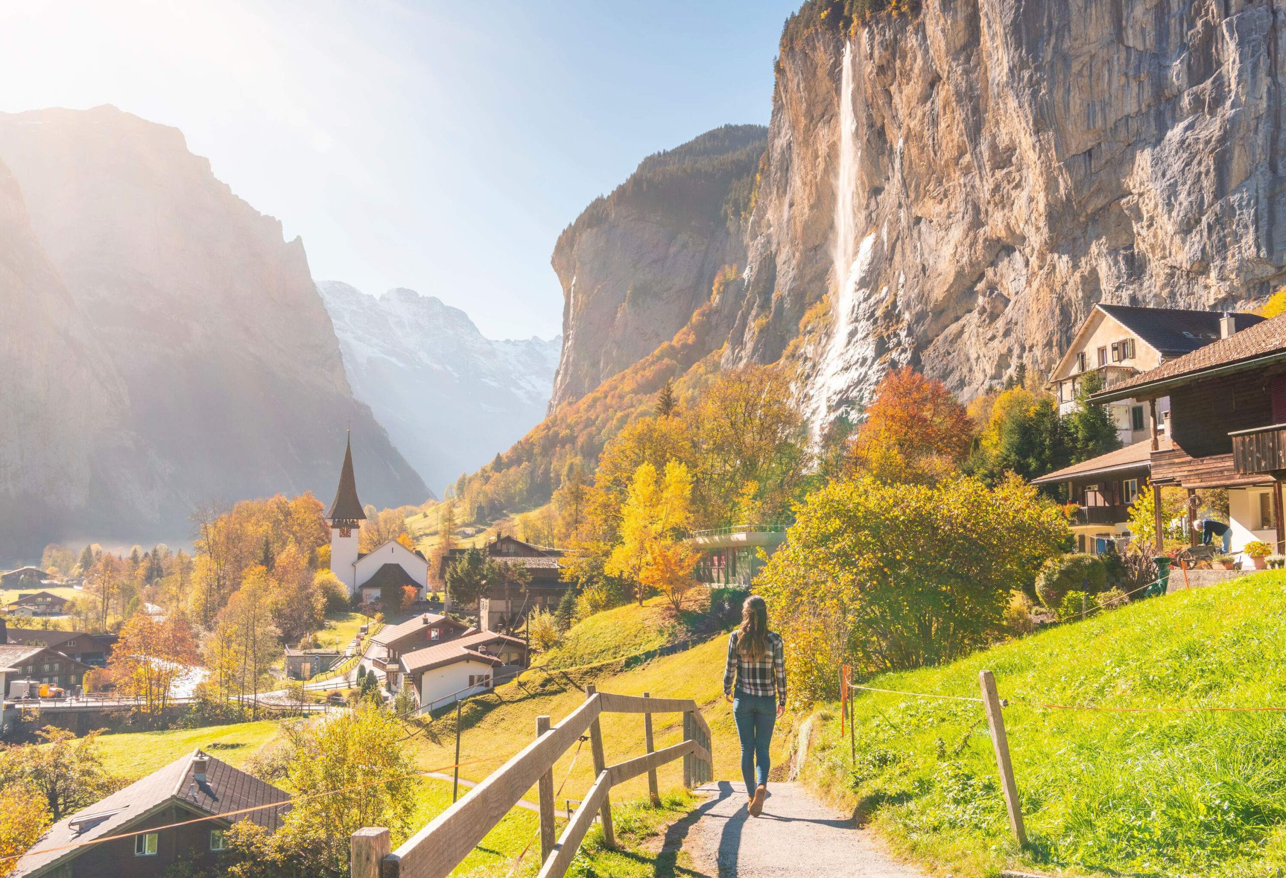 A person walks along a scenic path through a picturesque village nestled among steep cliffs, basking in the warm sunlight and taking in the breathtaking natural beauty of their surroundings.