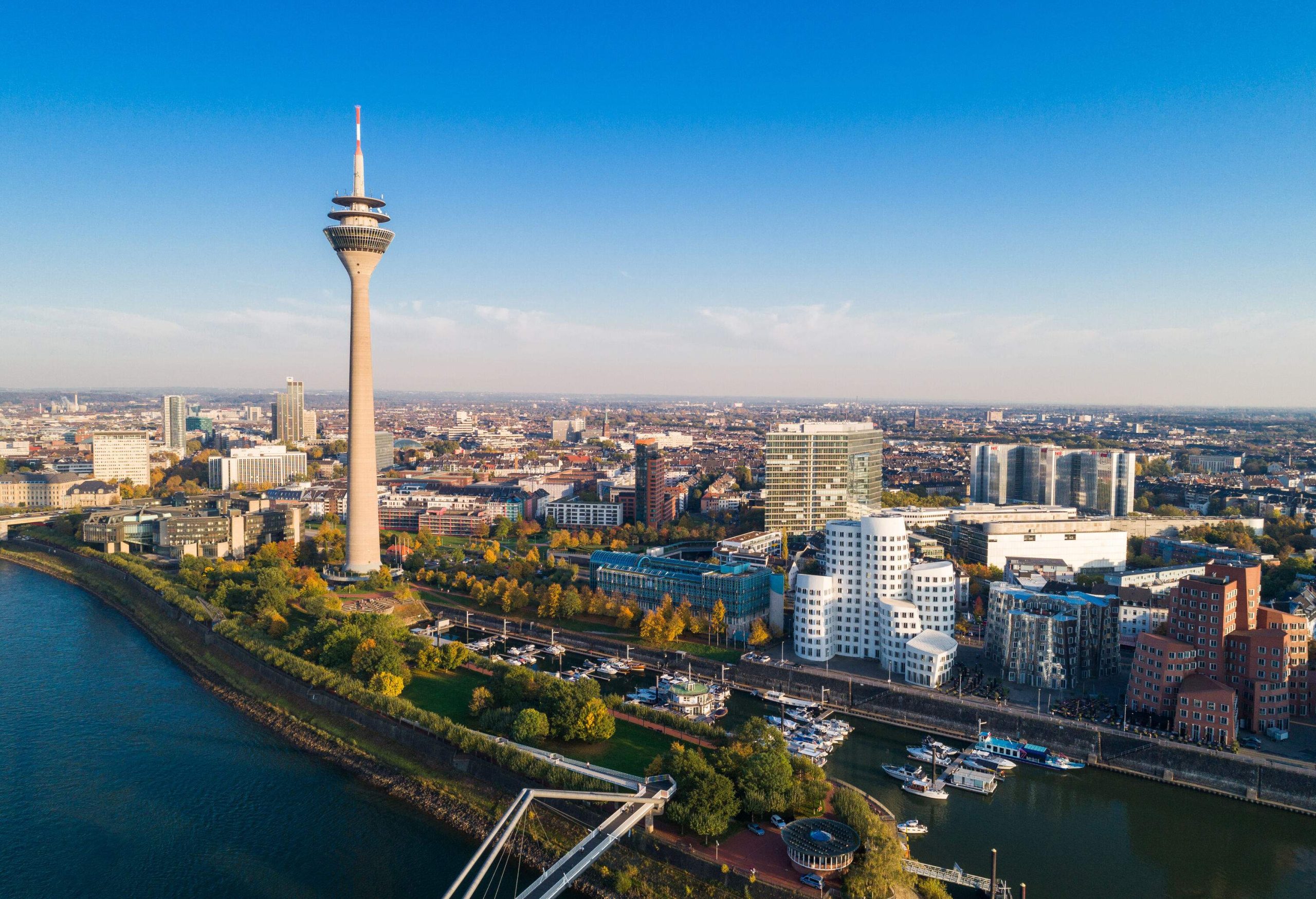 Rheinturm is a telecommunications tower overlooking the river, anchored boats in a marina, and the urban landscape of a city.