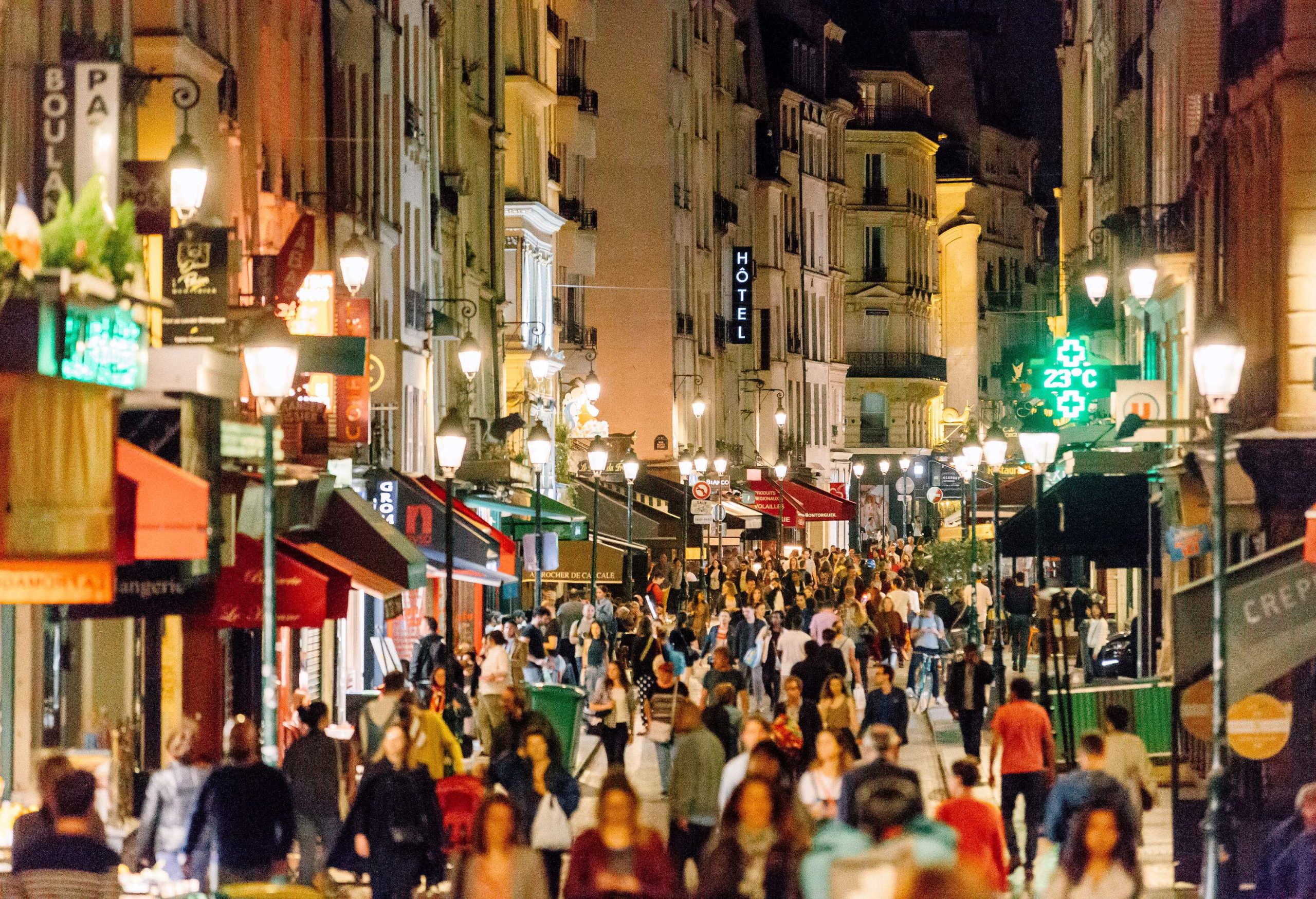 A night-time scene of a crowded street lined with lit street lamps, stores, and restaurants.