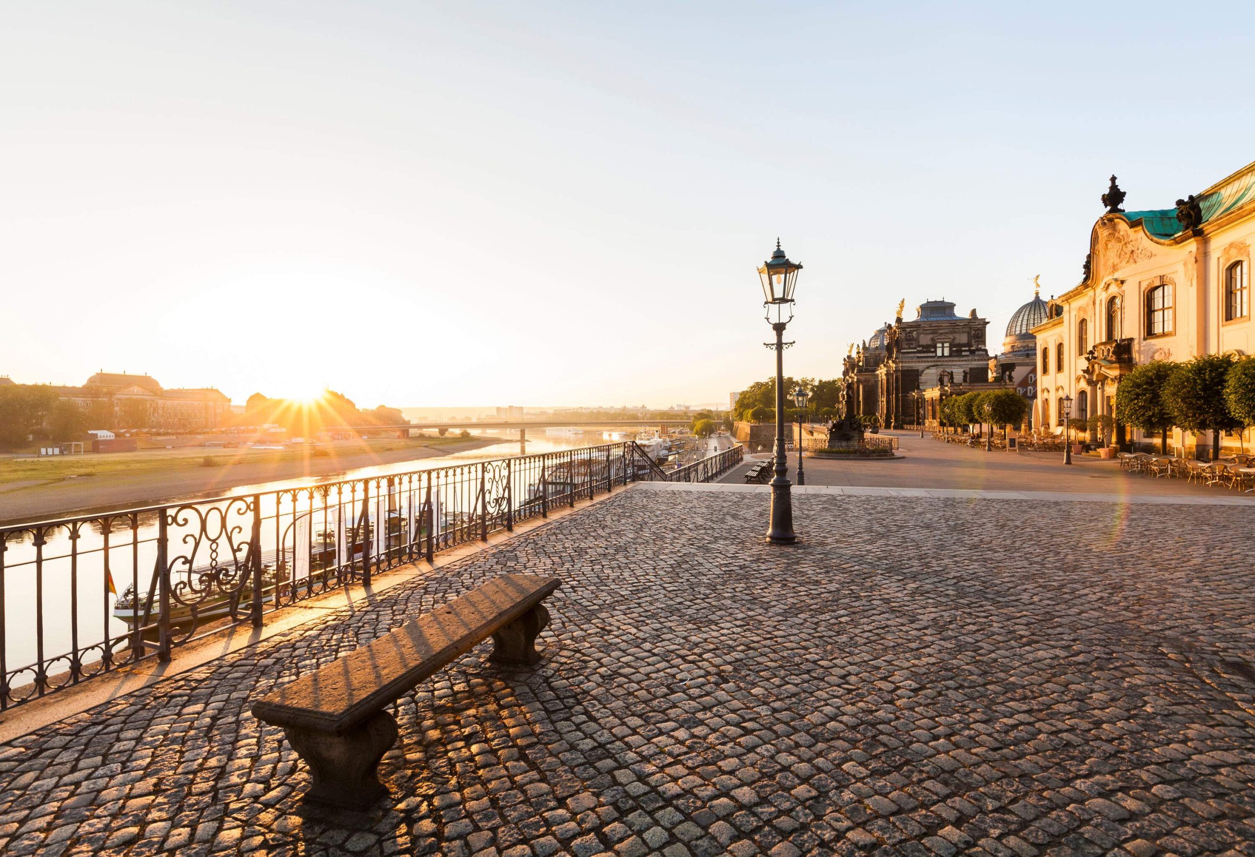 A broad cobbled promenade guarded with wrought irons, scattered trees, light posts, and tall buildings alongside a river.