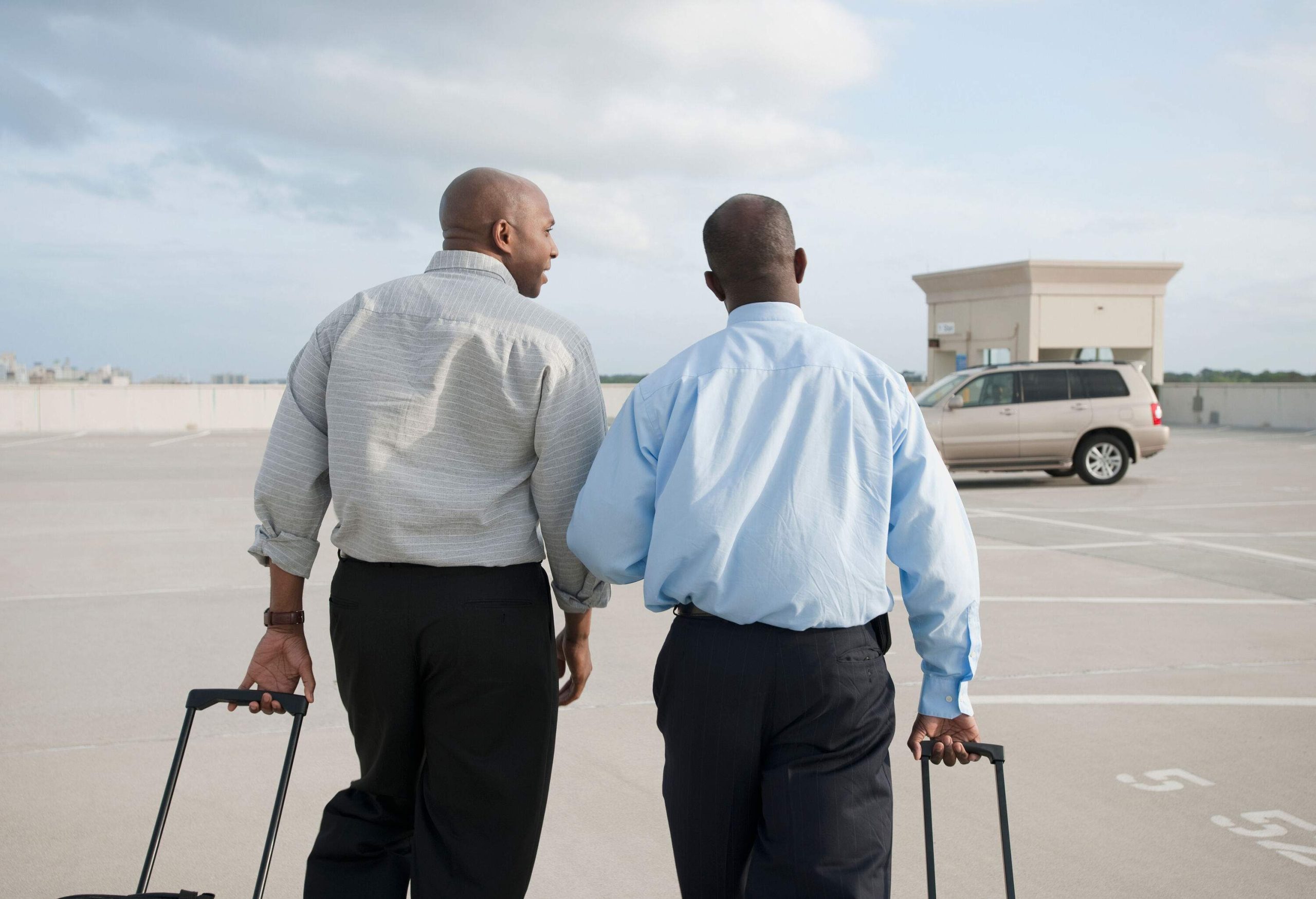 Two individuals, dressed in black pants and long sleeves, drag their luggage across a parking lot as they approach a waiting car.