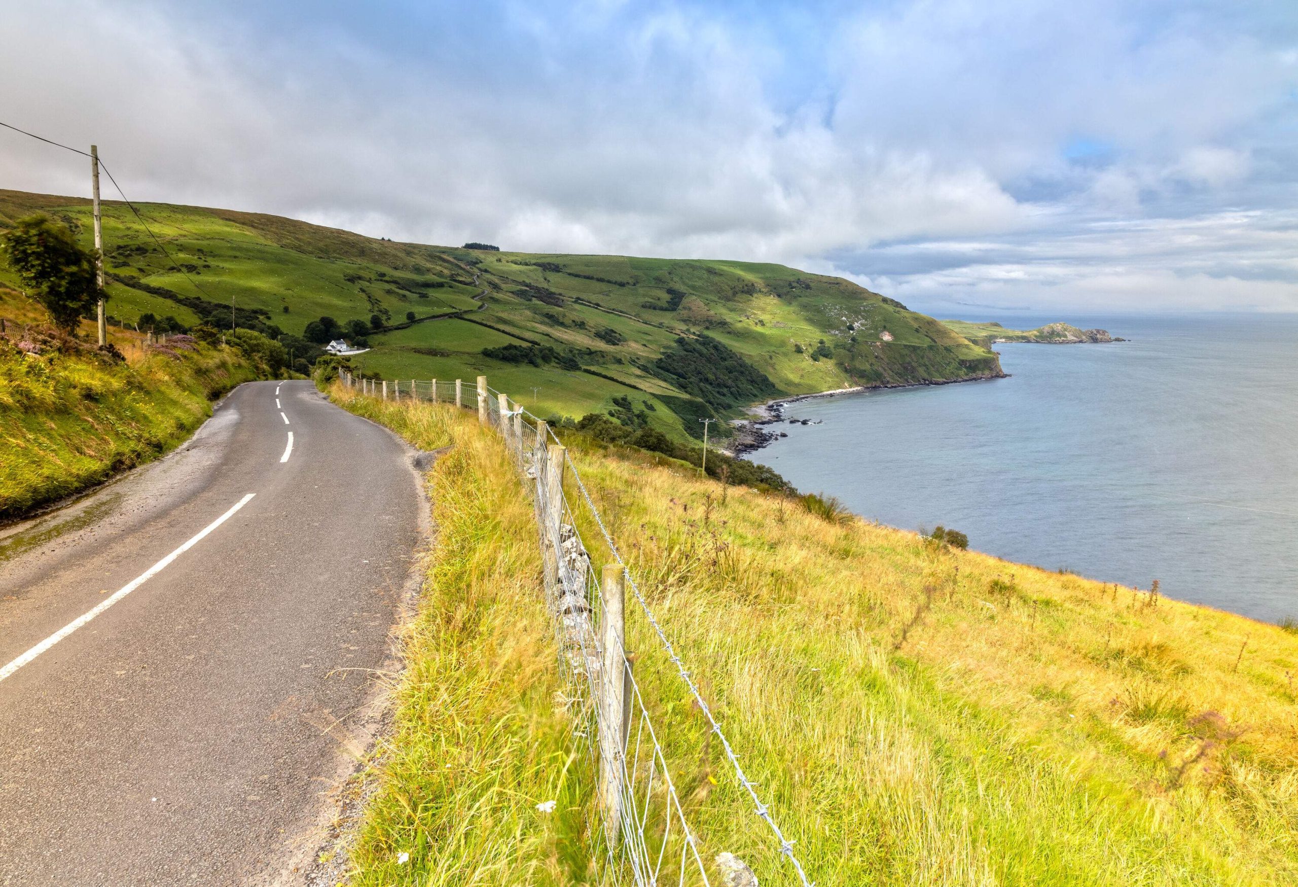 A hillside roadway by the sea with a barbed-wire fence along the grass field.