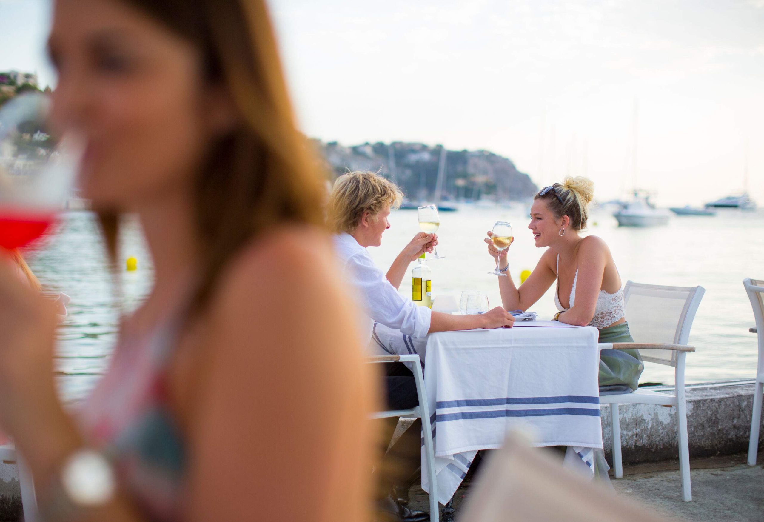 A blurred image of a woman with a background of a happy couple enjoying their drinks at a waterfront restaurant.