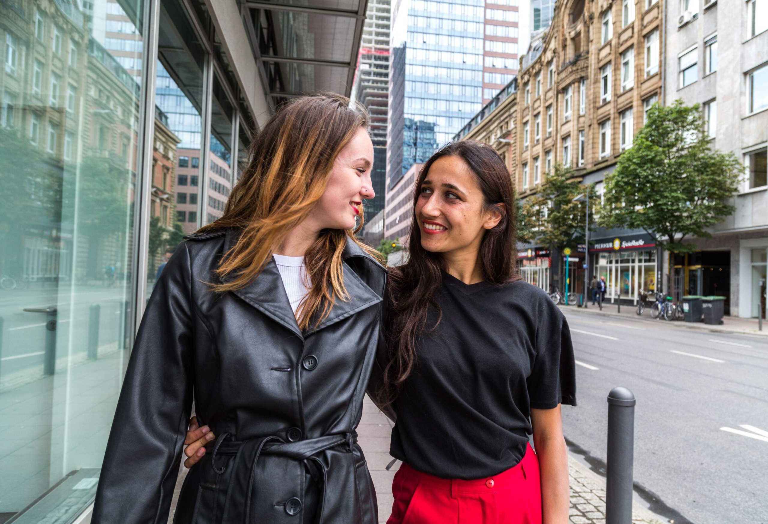 Two happy women in casual attire look at each other while standing on a street lined with classic and modern buildings.