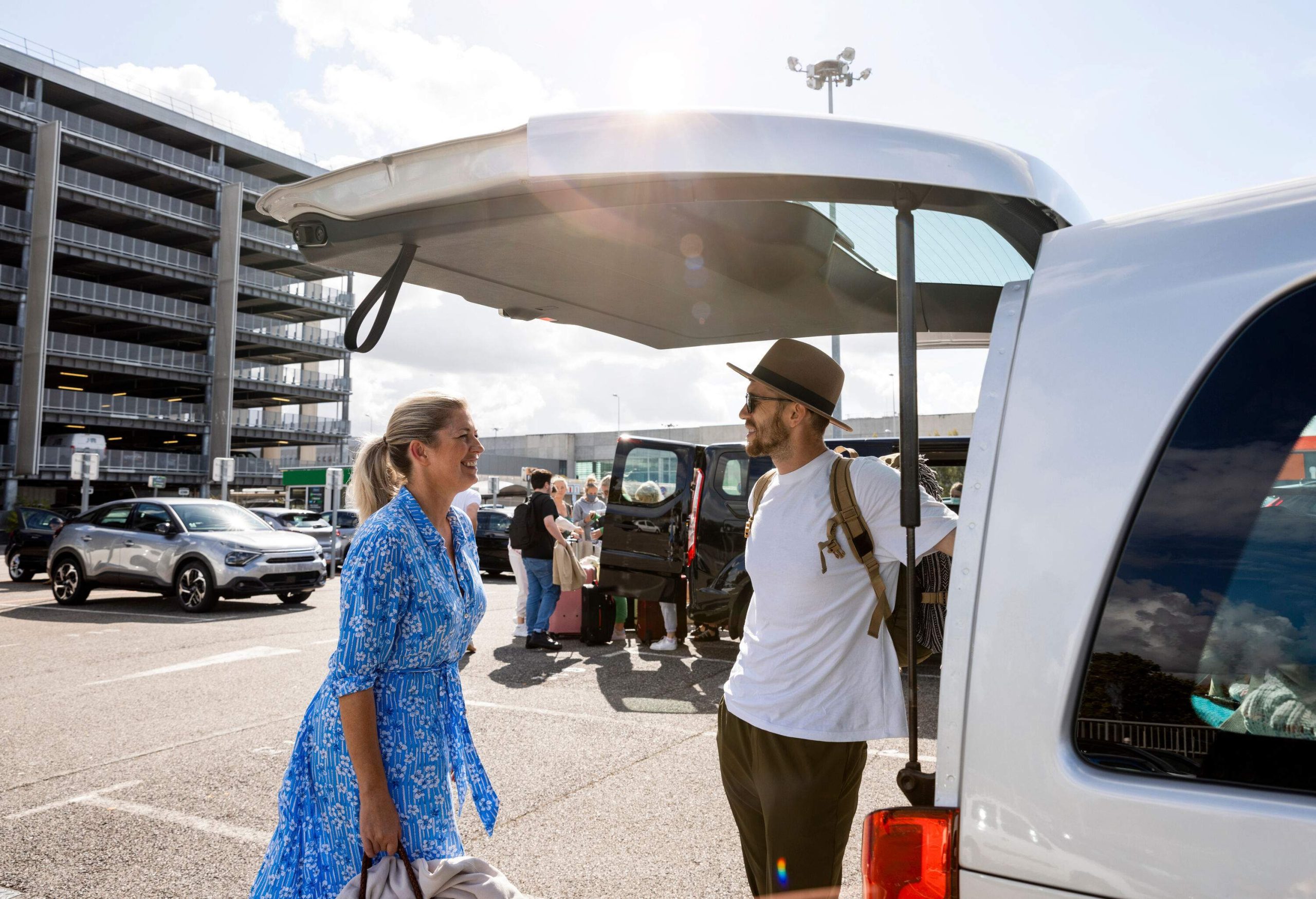 Two individuals engaged in a lively conversation at the back of a white van parked in a spacious parking lot.