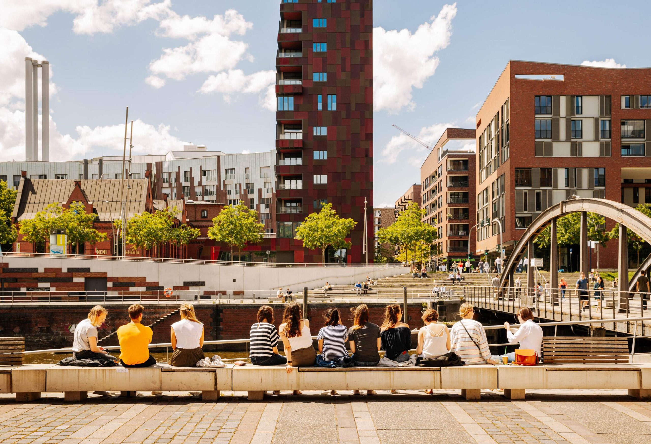 People leisurely sit along a long bench on a river promenade, enjoying the scenic view of buildings and trees across the river.