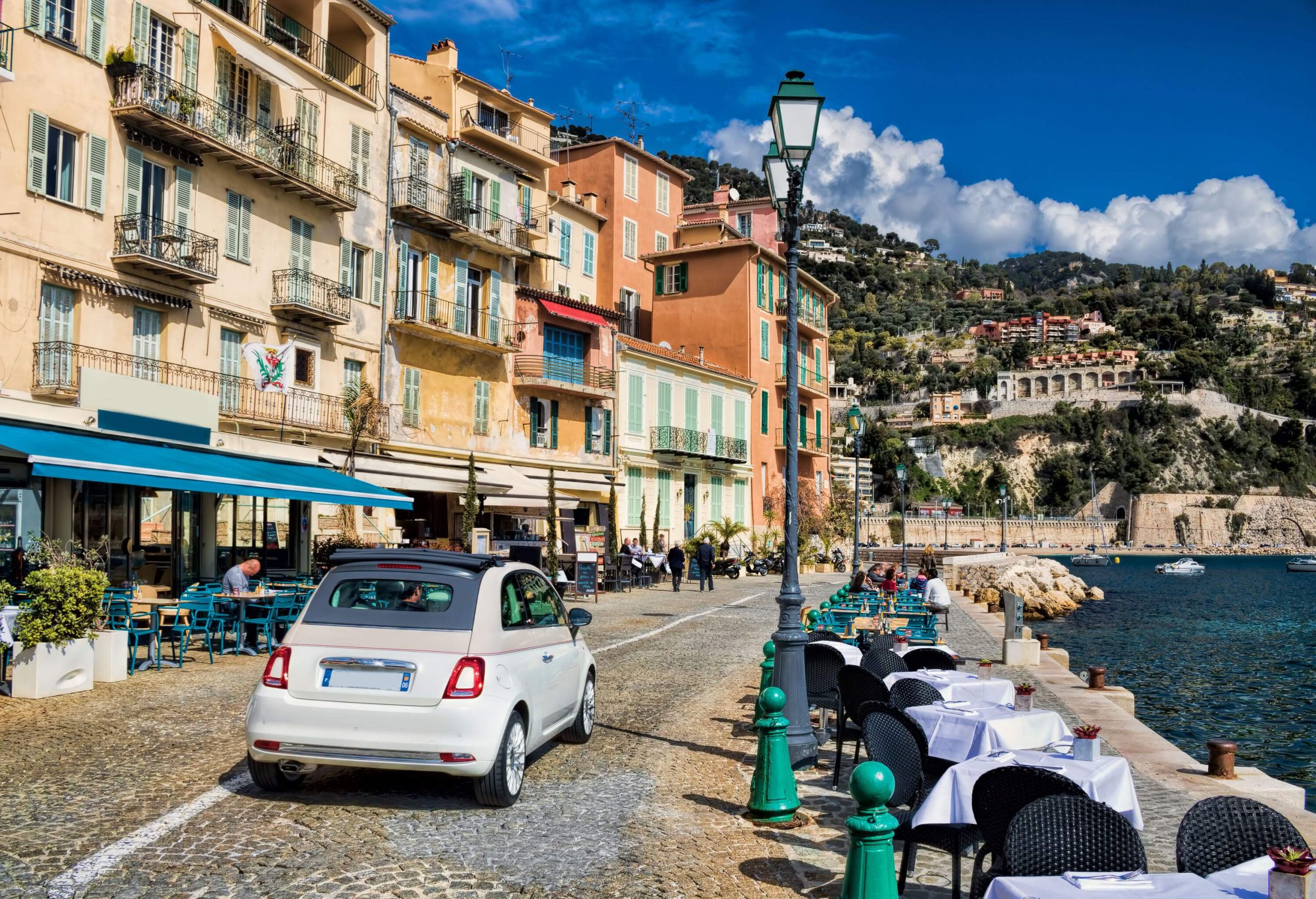 A roadway lined with dinner tables and outdoor cafes in row buildings that face a body of water are backed by a building-covered slope.