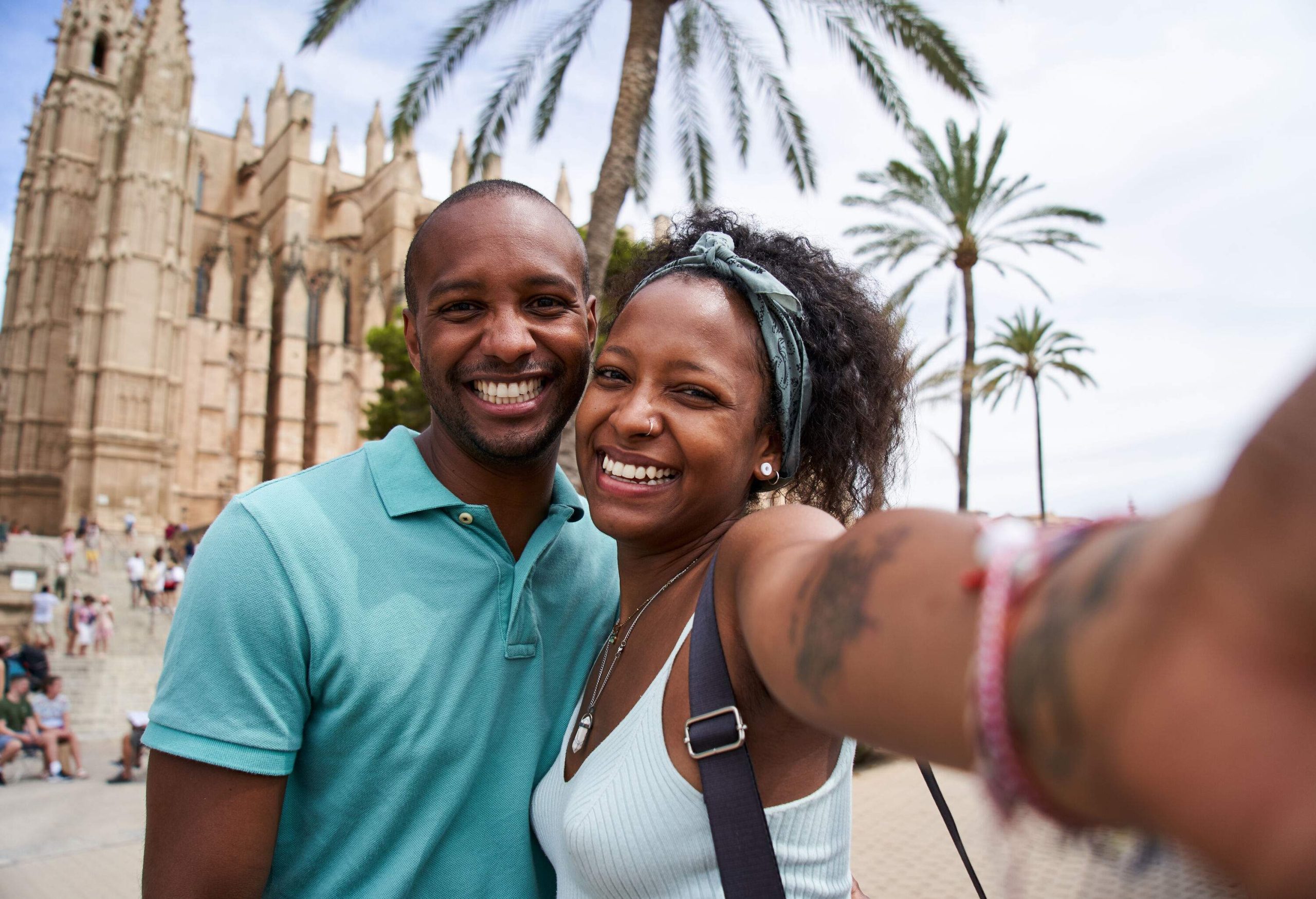 A young African American couple is taking a selfie with an out-of-focus cathedral in the background that has numerous bystanders in front.