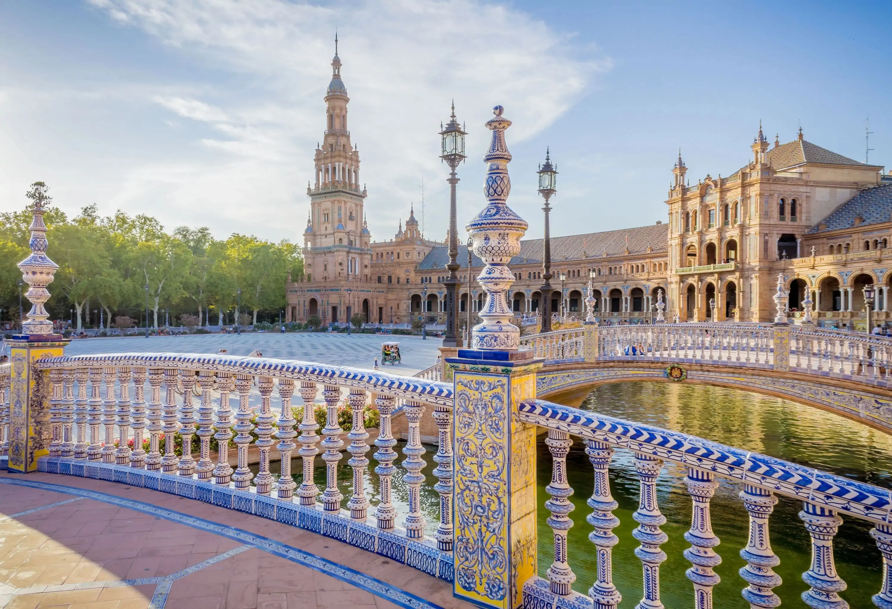 A balcony with ornate railings overlooks a river bridge and a public square beside a building with a pointed tower.