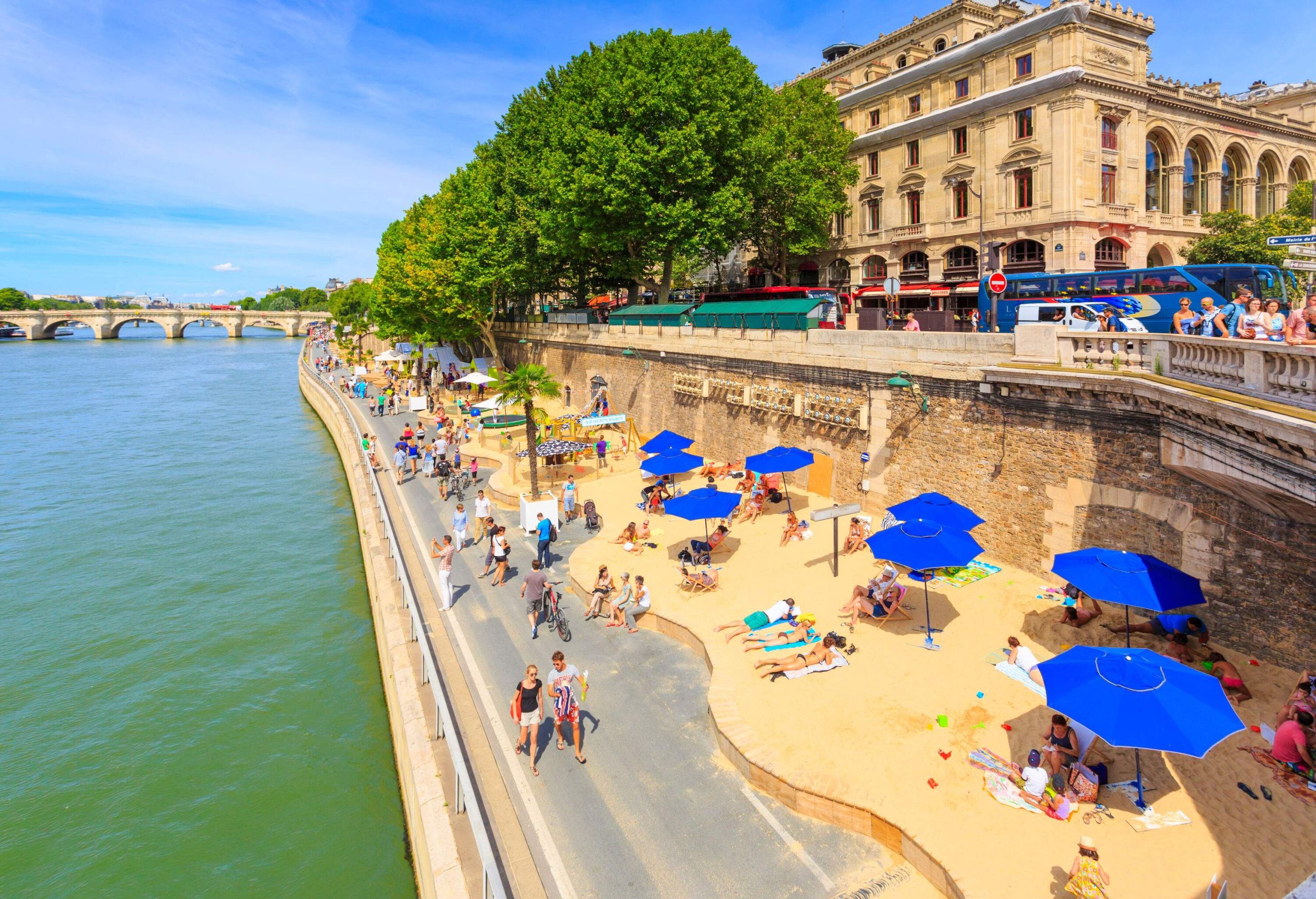 A riverside promenade where people walk contains a sand platform where people relax under parasols.