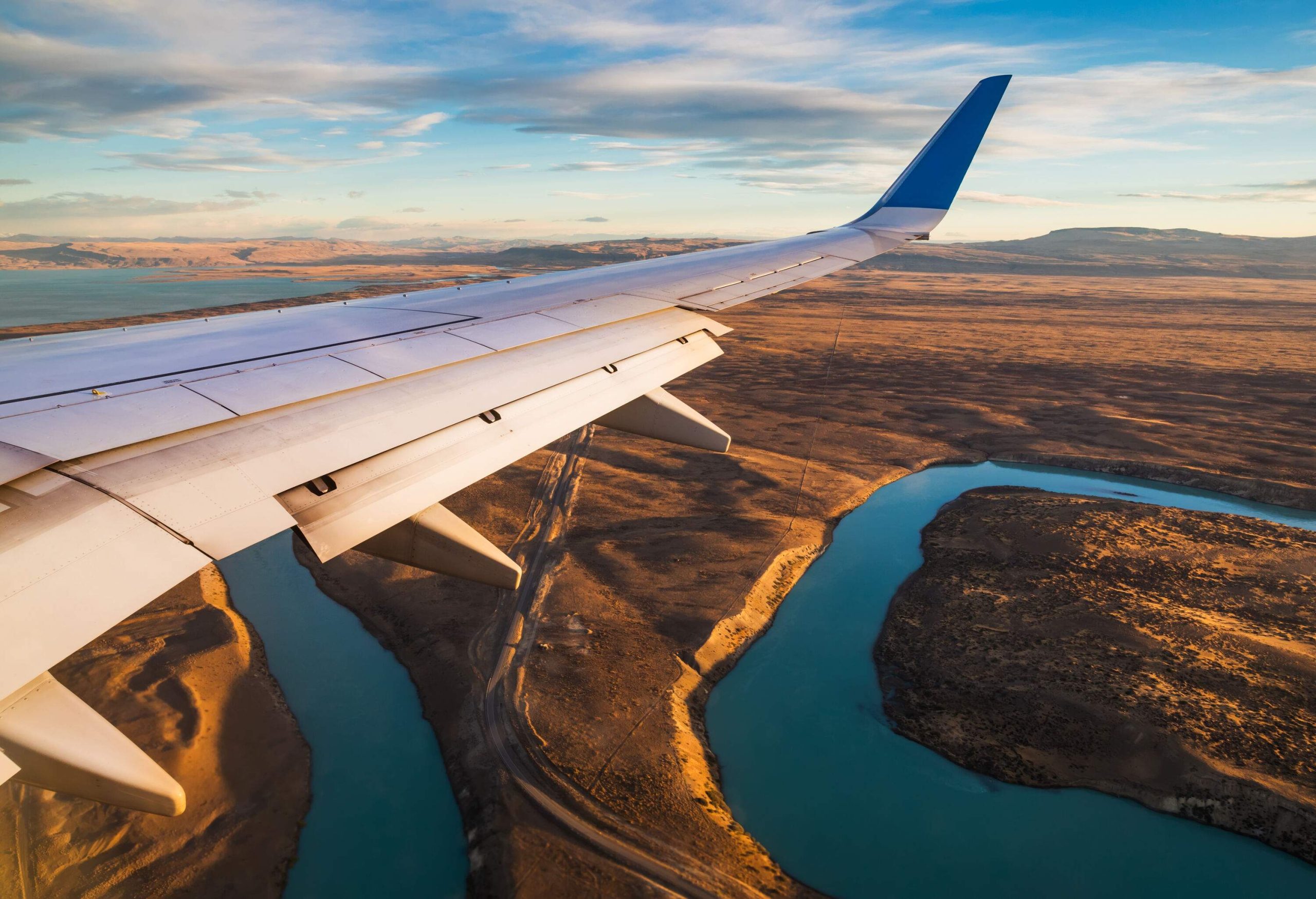 The sleek wing of a plane soars over a vast expanse of sandy terrain, with a winding river cutting through the landscape below.