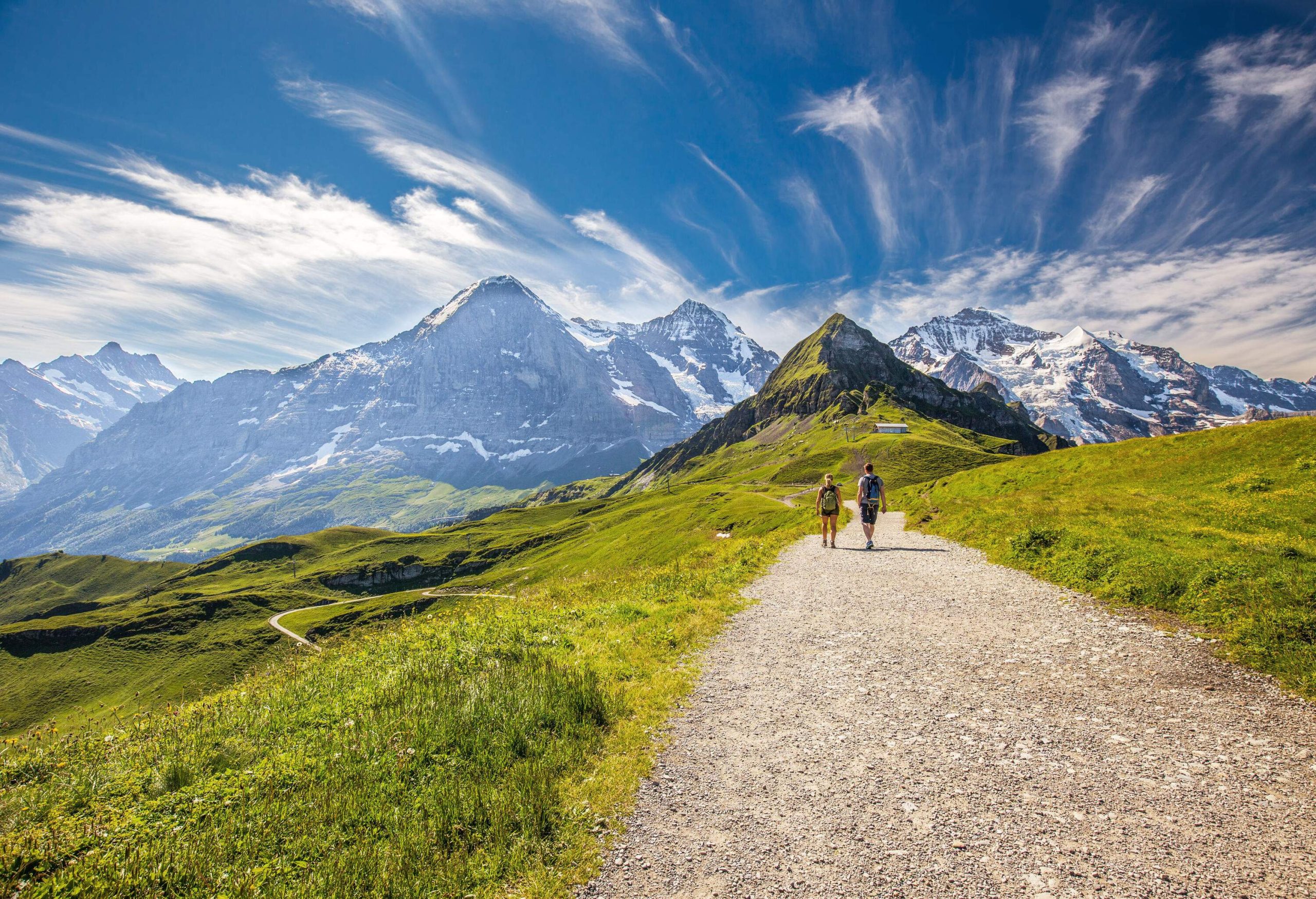 Two people walk along a sandy path surrounded by greenery, with snow-capped crags and streaks of clouds in the sky.