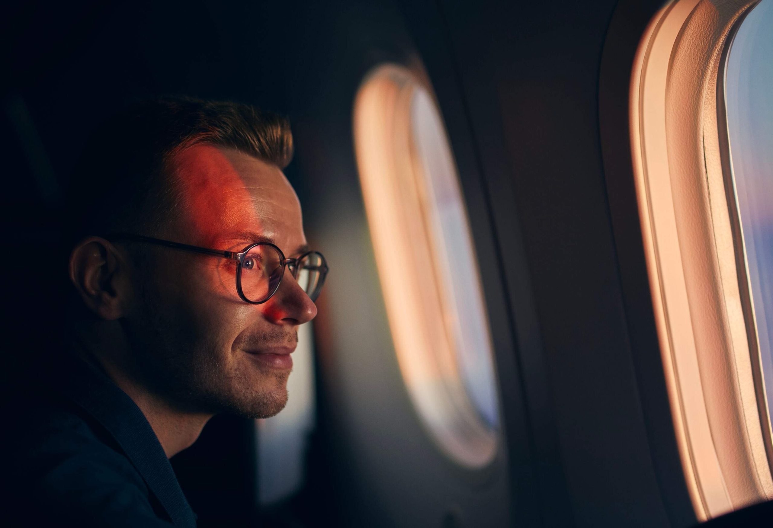 A man wearing eyeglasses smiling as he looks out the window of an airplane.