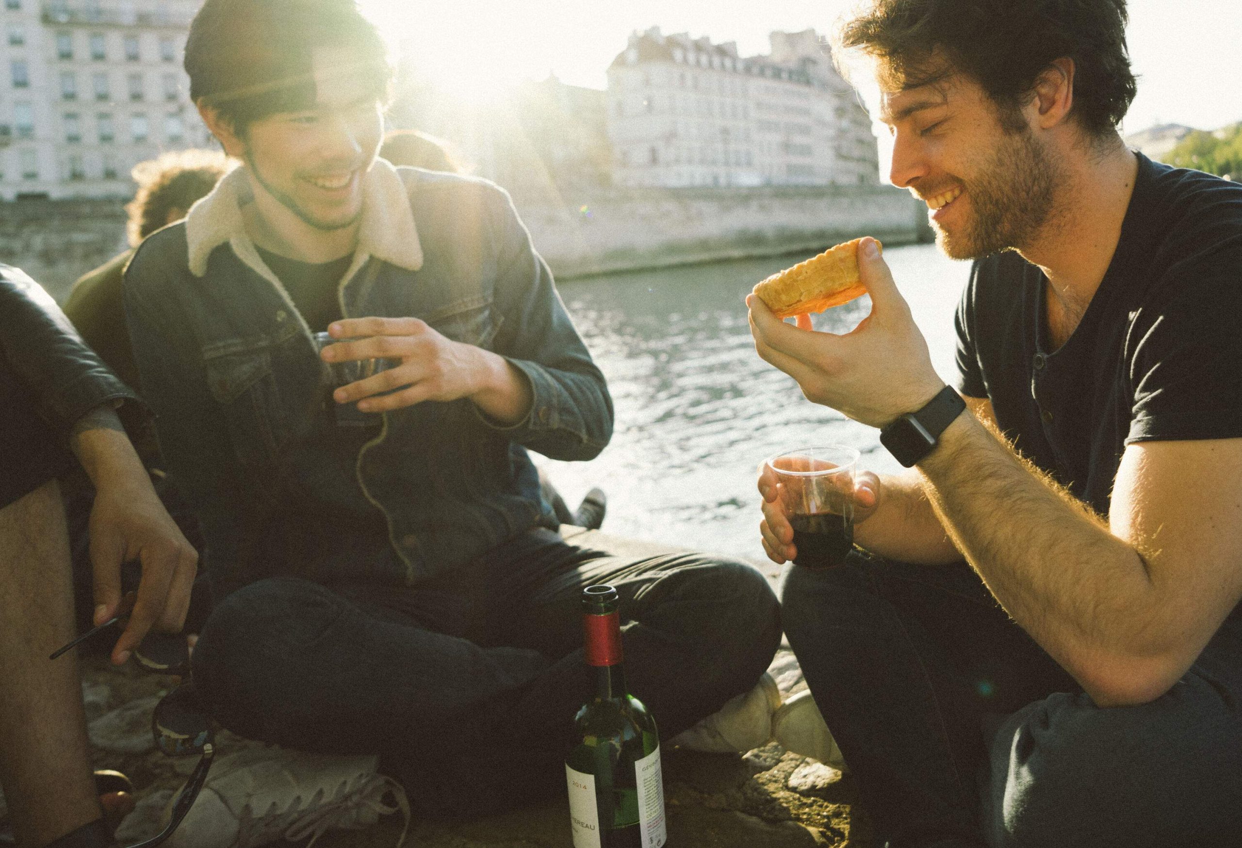 Two men enjoying food and wine by the river as the sun shines through the buildings behind them.