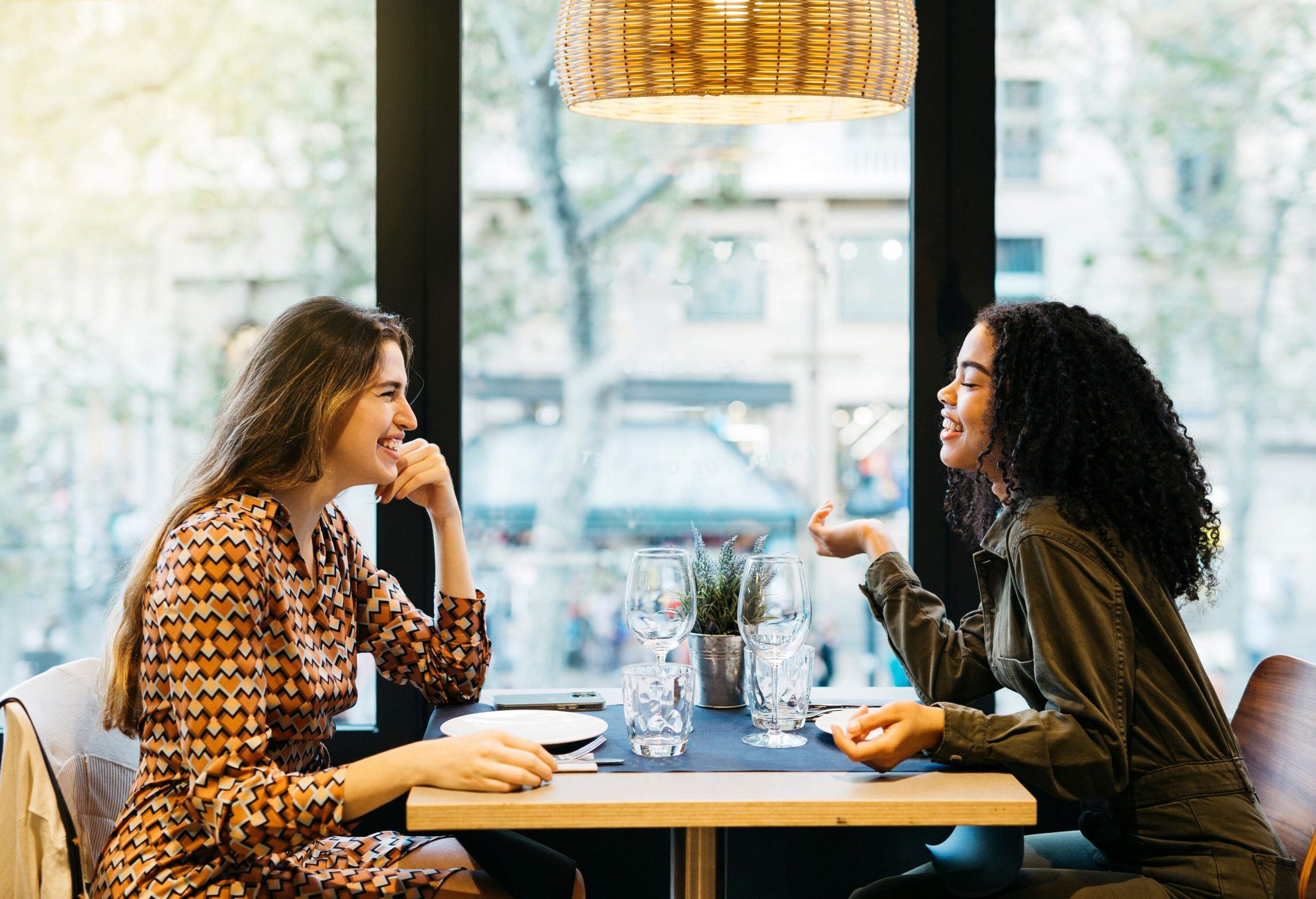 Two female friends in casual attire sit on a table while having a conversation.