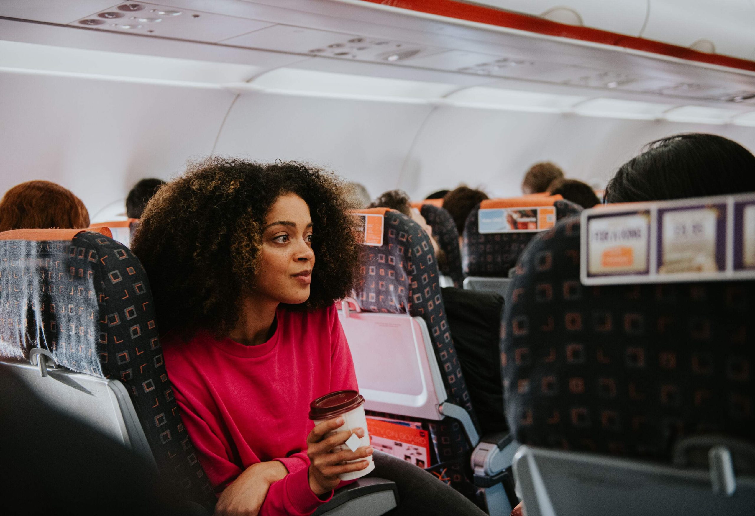 A young woman sits on a crowded plane on an aisle seat. She clutches a cup of coffee and looks perplexed and thoughtful.