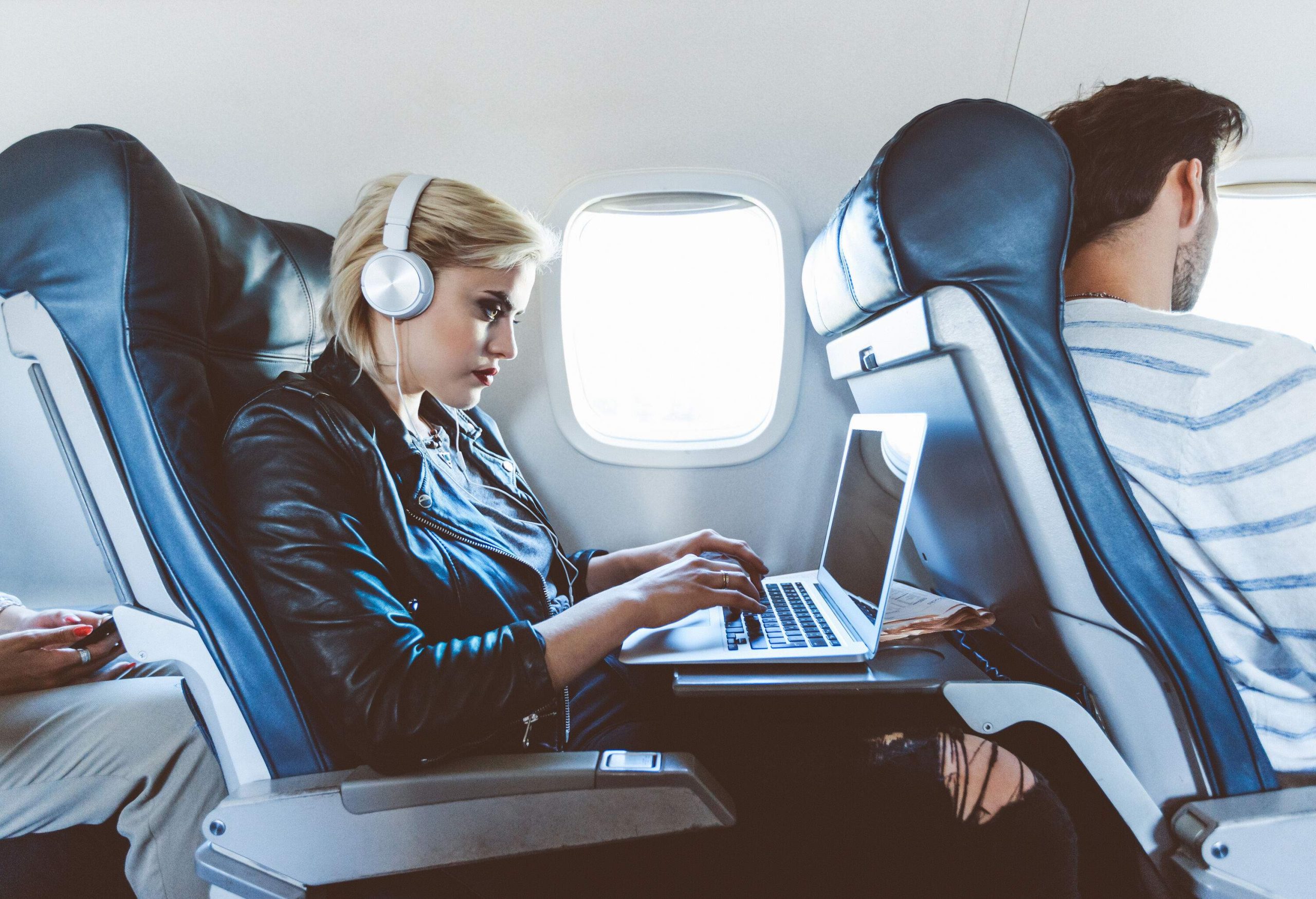 A female traveller on a flight, engrossed in her work, using a laptop computer, and wearing headphones.