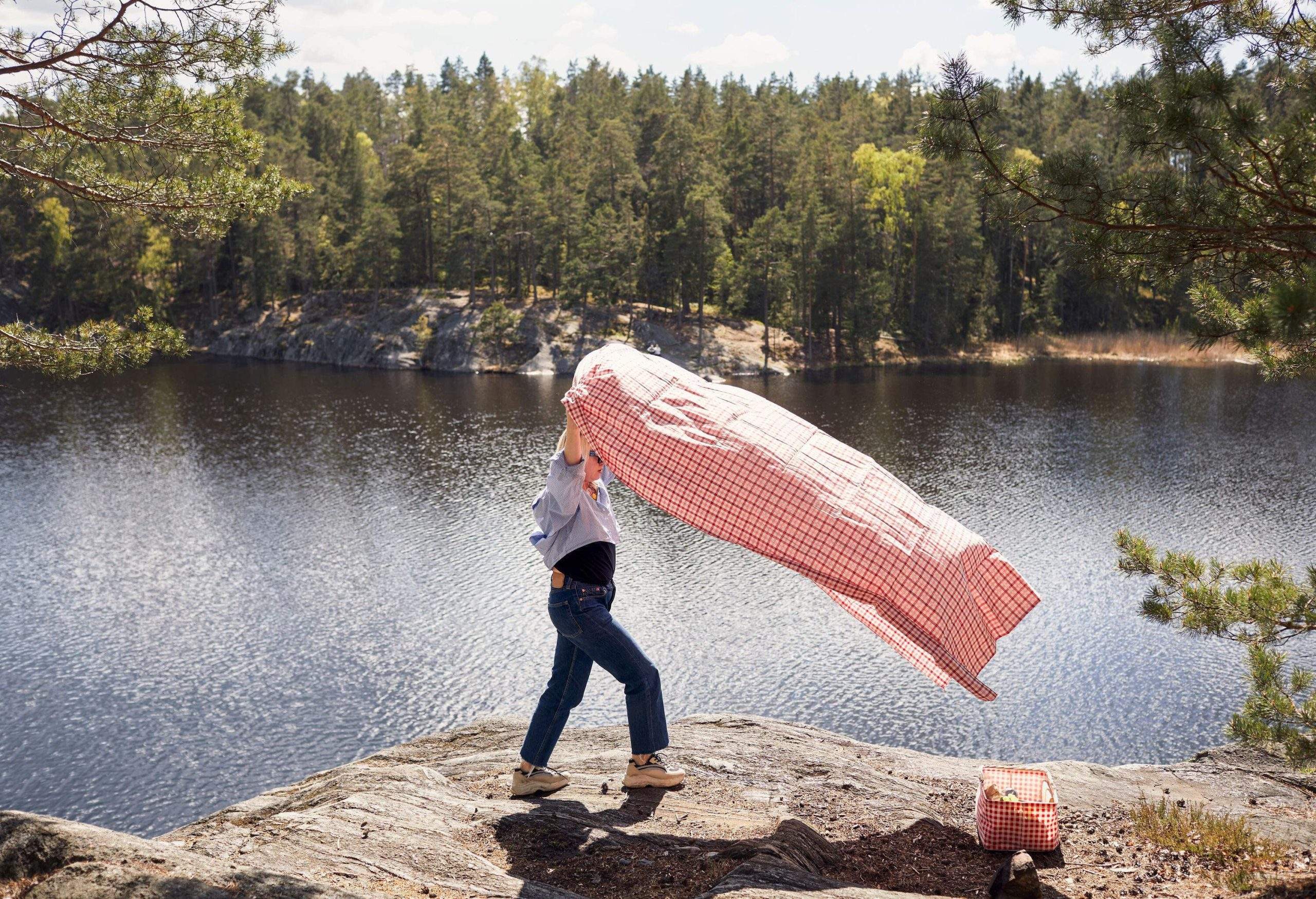 A woman with a pink picnic blanket on a cliff by the lake surrounded by tall green trees.