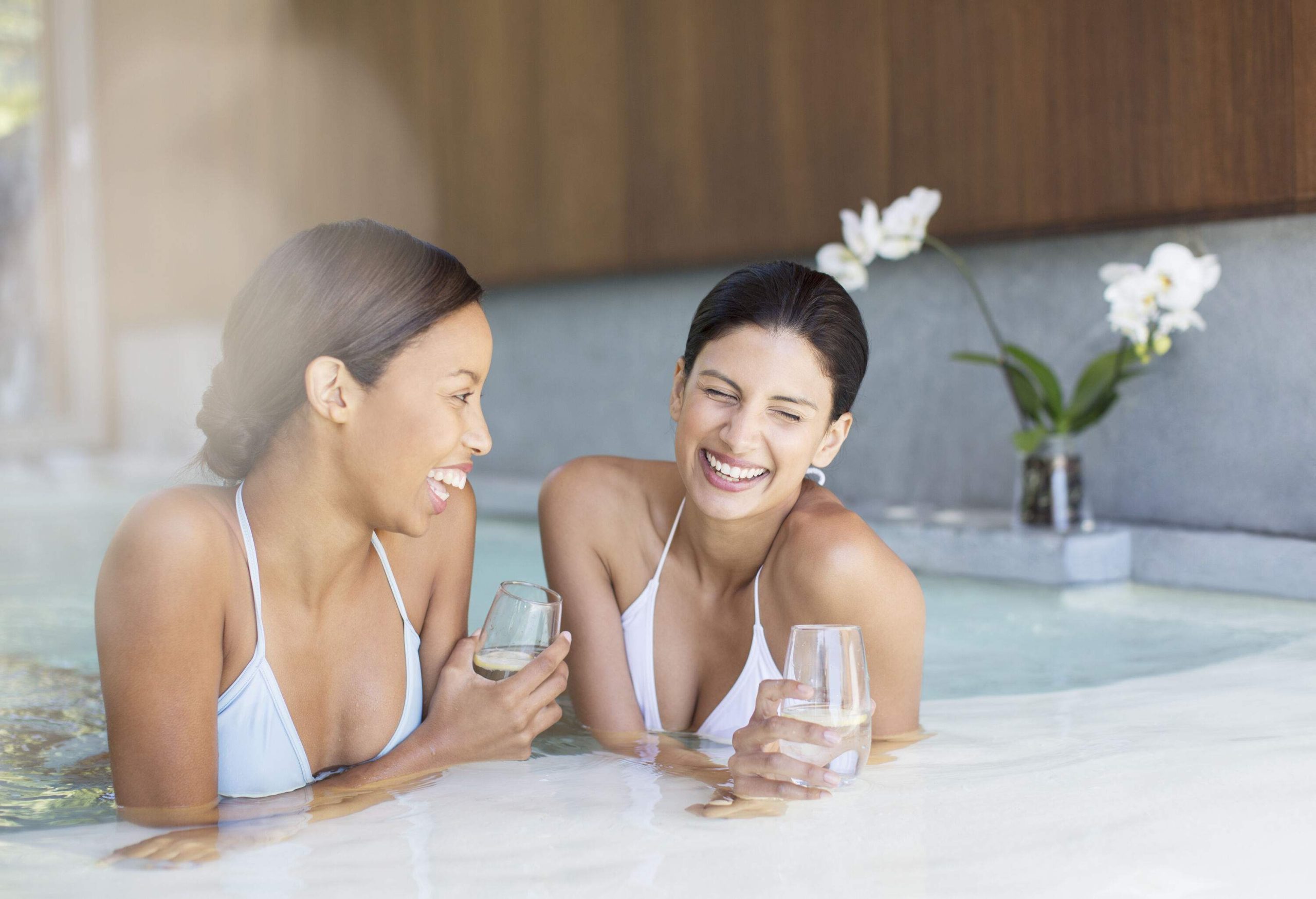 Two happy women in white swimsuits are submerged in a steaming hot tub while holding a glass of white wine.