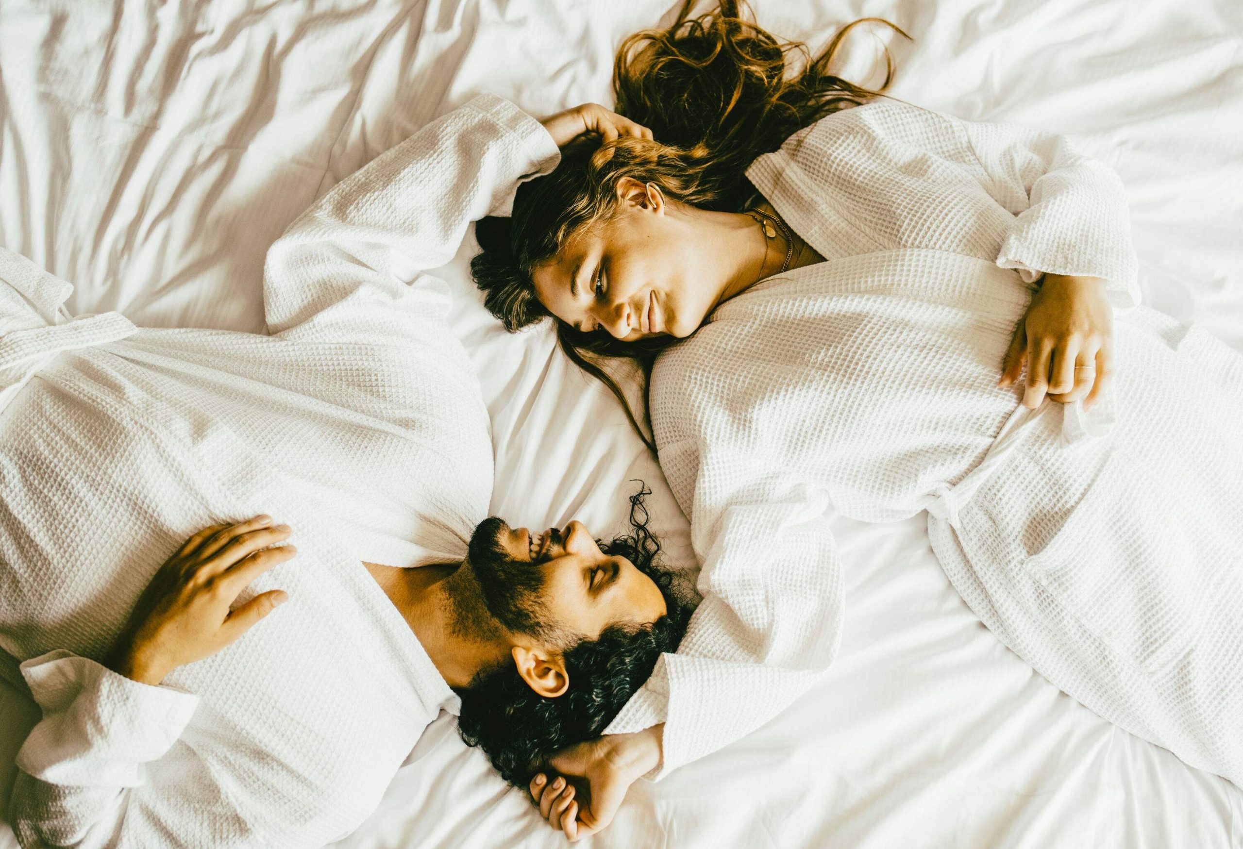 A lovely couple in white robes lying head to head in the bed with their feet in opposite directions.