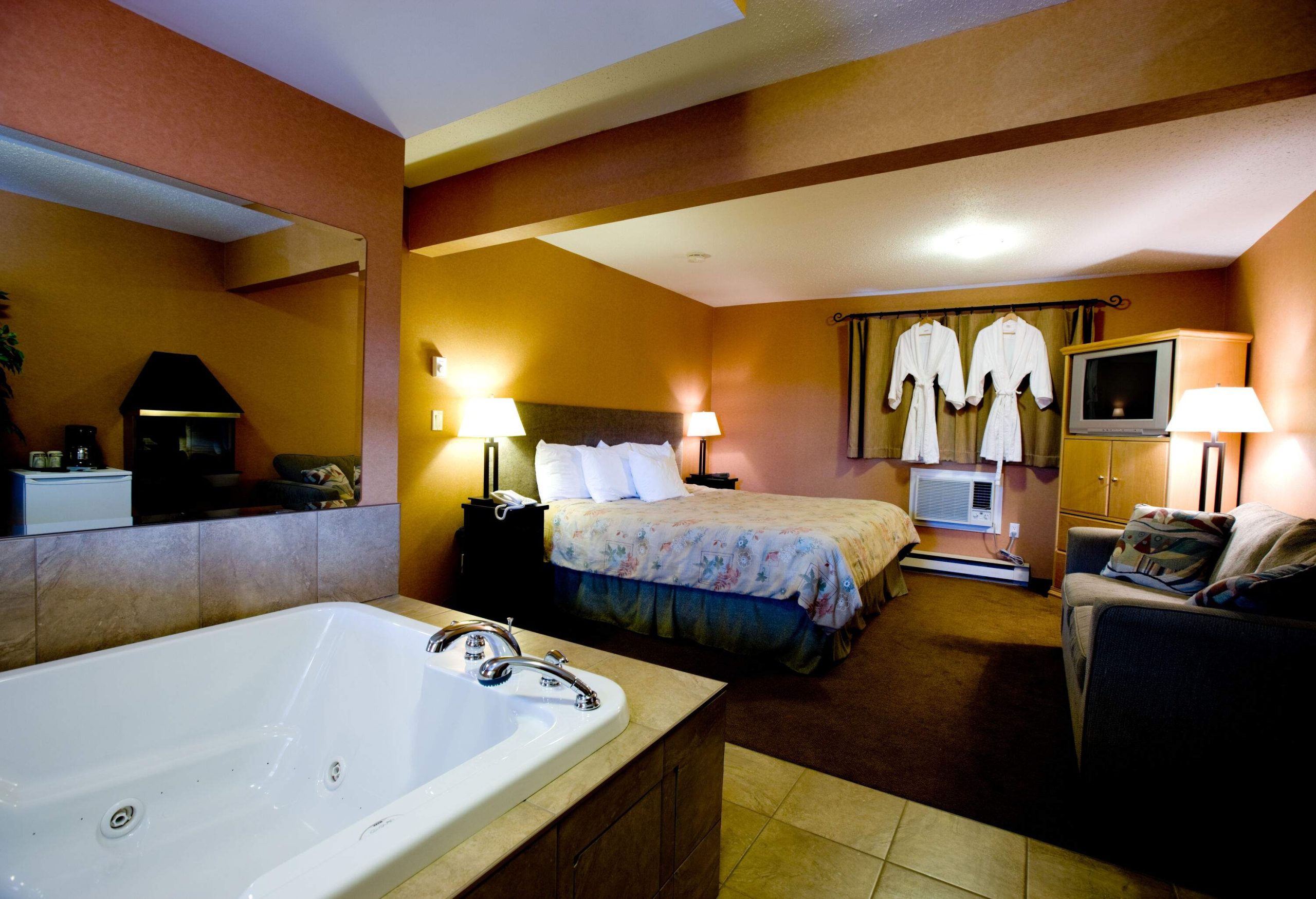 A hotel room with a cosy bed, a sofa and a jacuzzi with two bathrobes hanging on the window curtain.