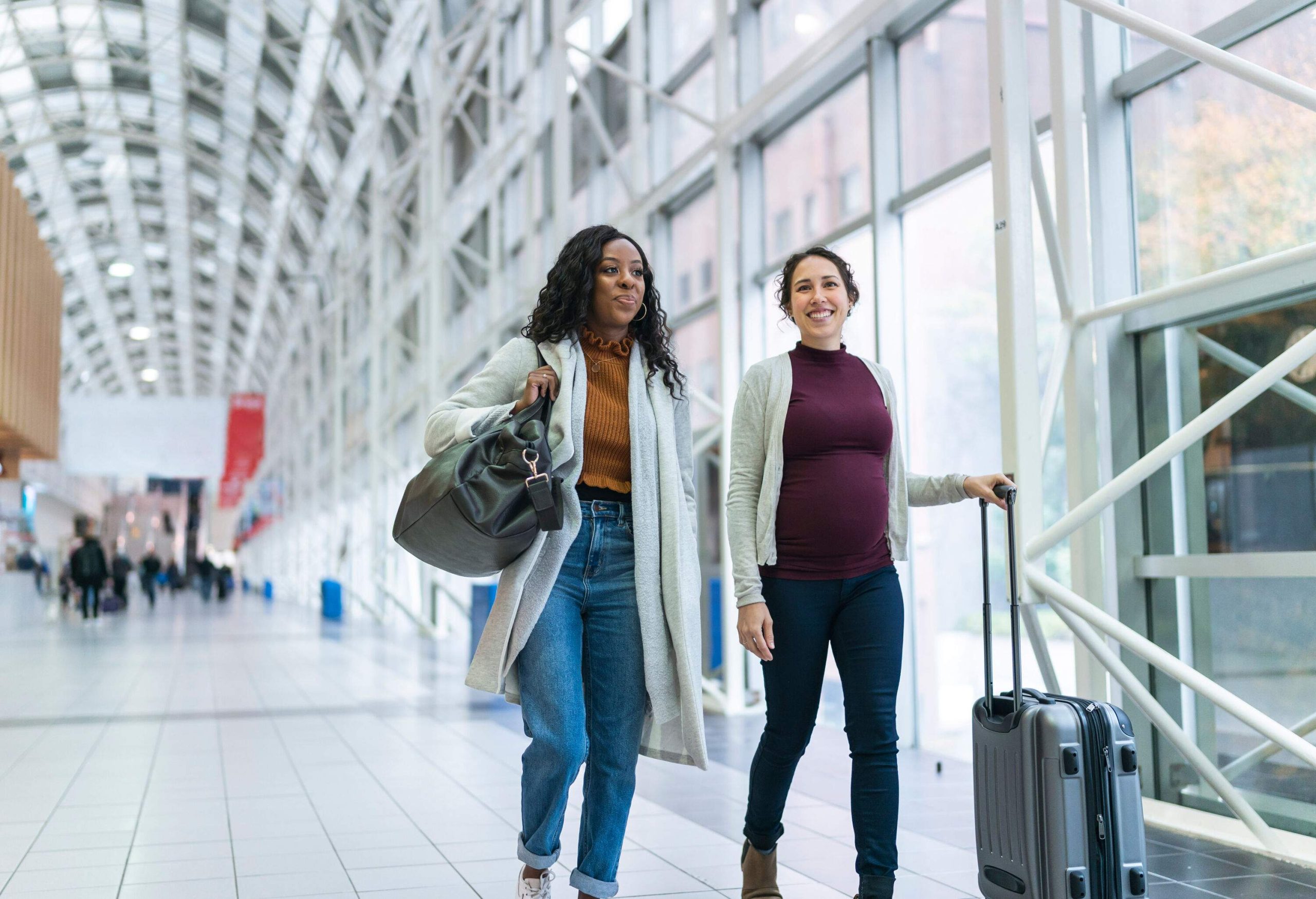 Two female friends with bright smiles were walking side by side through the airport, each carrying their luggage.